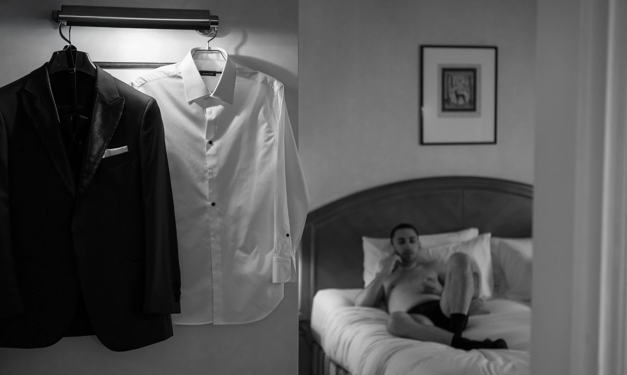 A black-and-white photo of a hotel room. In the foreground, a wardrobe displays a black suit jacket and a white dress shirt hanging side by side. In the background, a man is lying on a bed with his head propped up on pillows, looking at his phone. Th