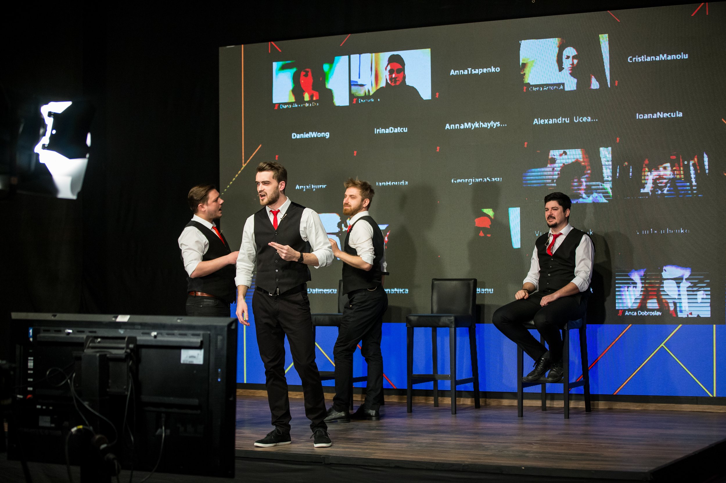 Three men dressed in formal vests and shirts speaking or engaging during a panel or presentation on a stage, with a large screen behind them displaying participant names and images.