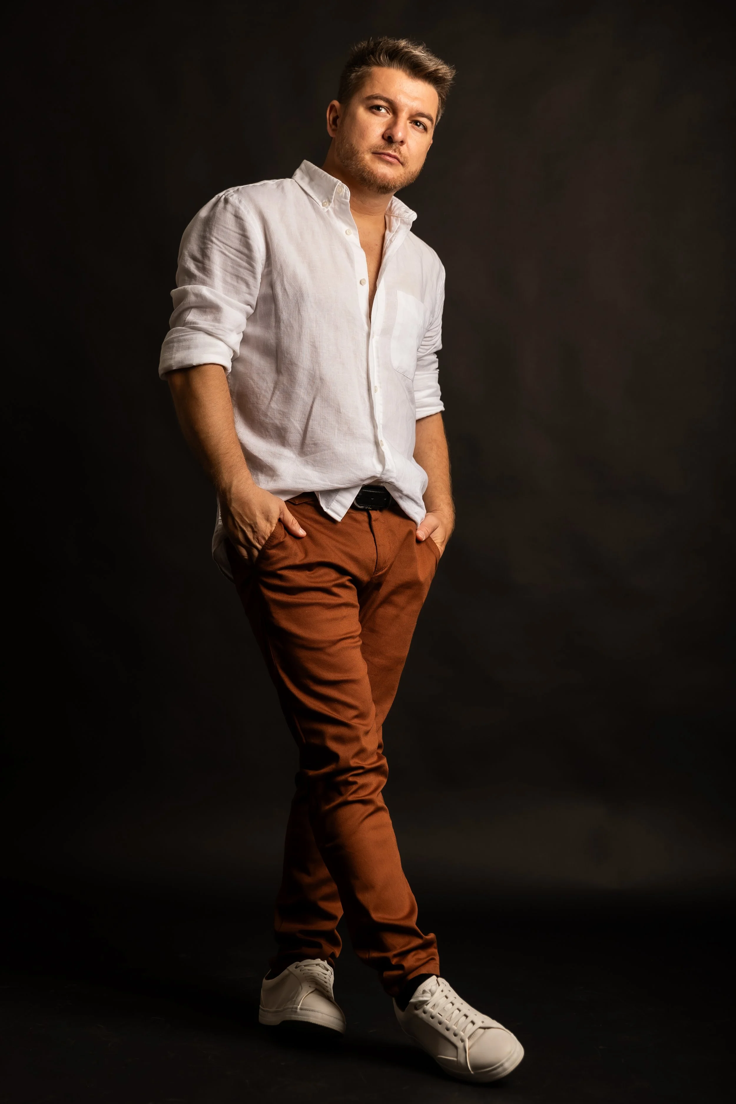 A young man with light brown hair and a beard wearing a white button-up shirt with rolled-up sleeves, brown pants, and white sneakers, standing against a dark background, looking confidently at the camera.