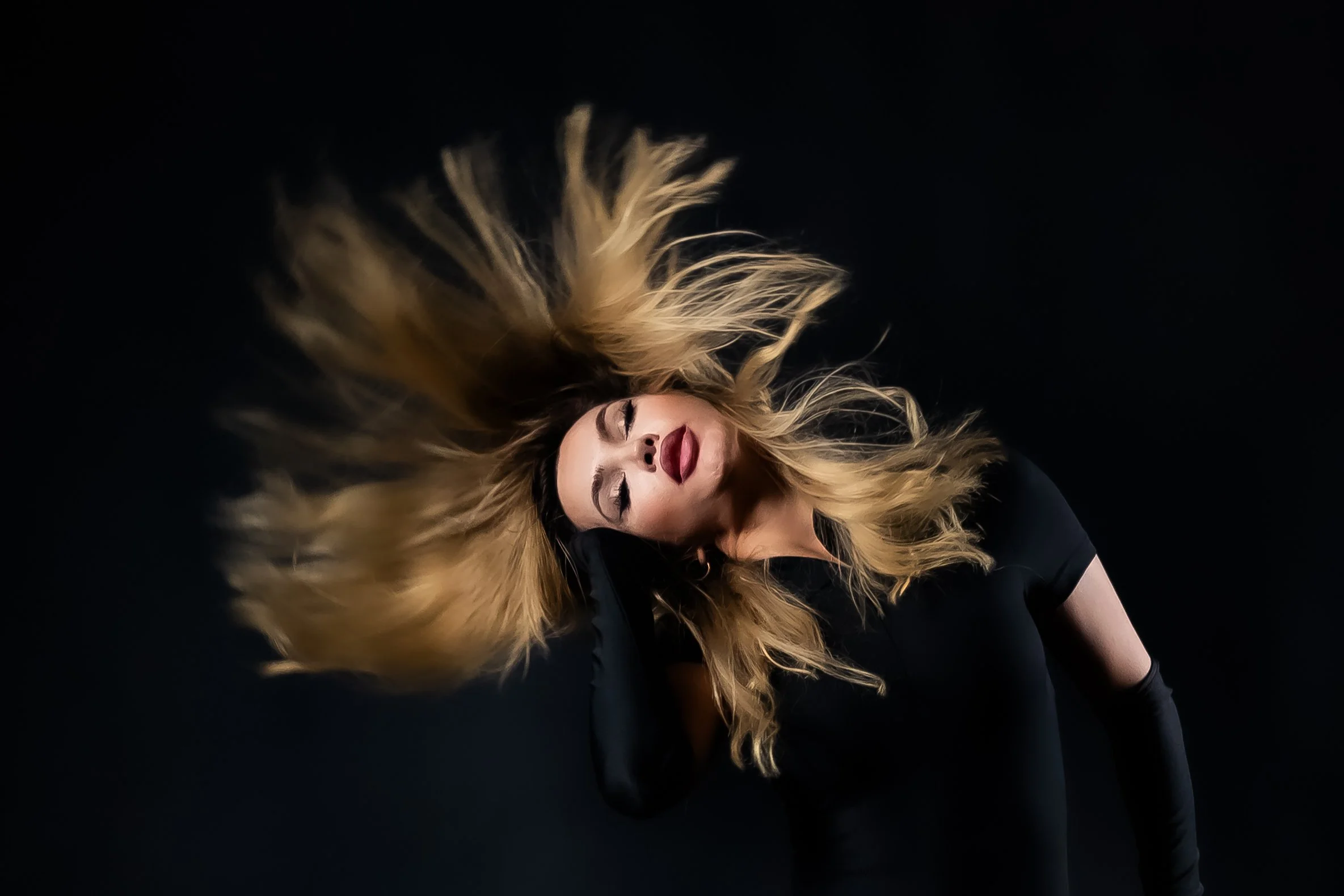 A woman with long, wavy blonde hair and makeup, wearing a black top, against a black background.