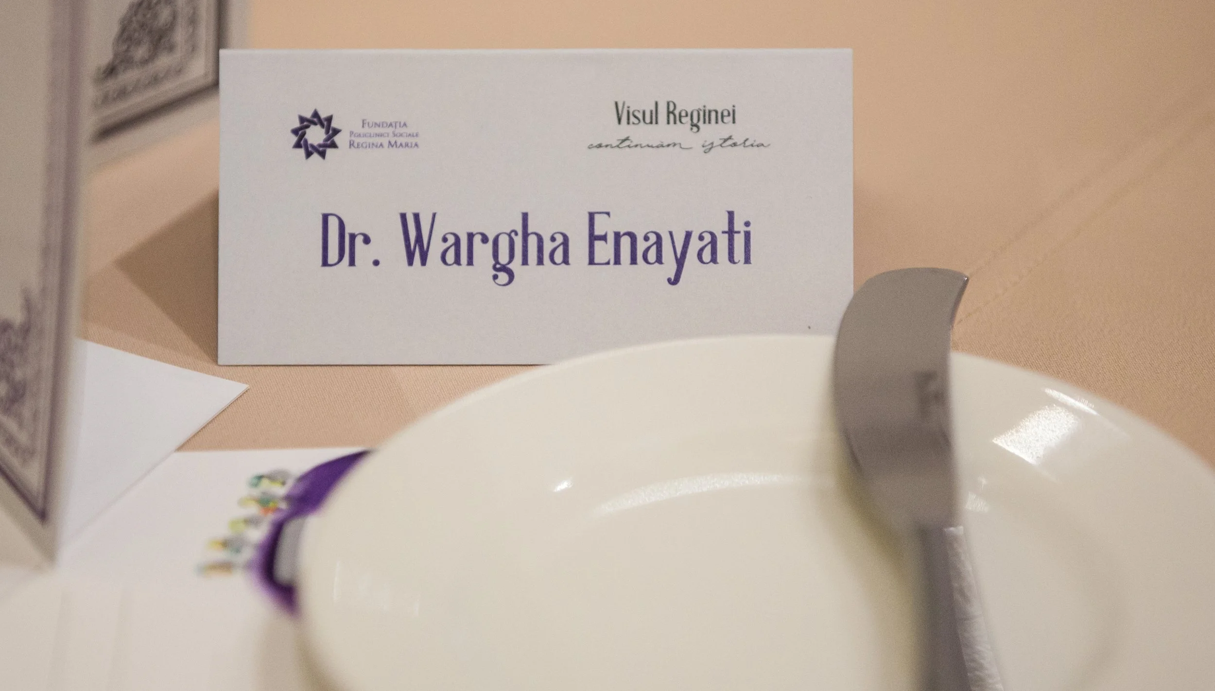 A nameplate with the text 'Dr. Wargha Enayati' on a table at an event, with a plate and some decorations partially visible in the foreground.