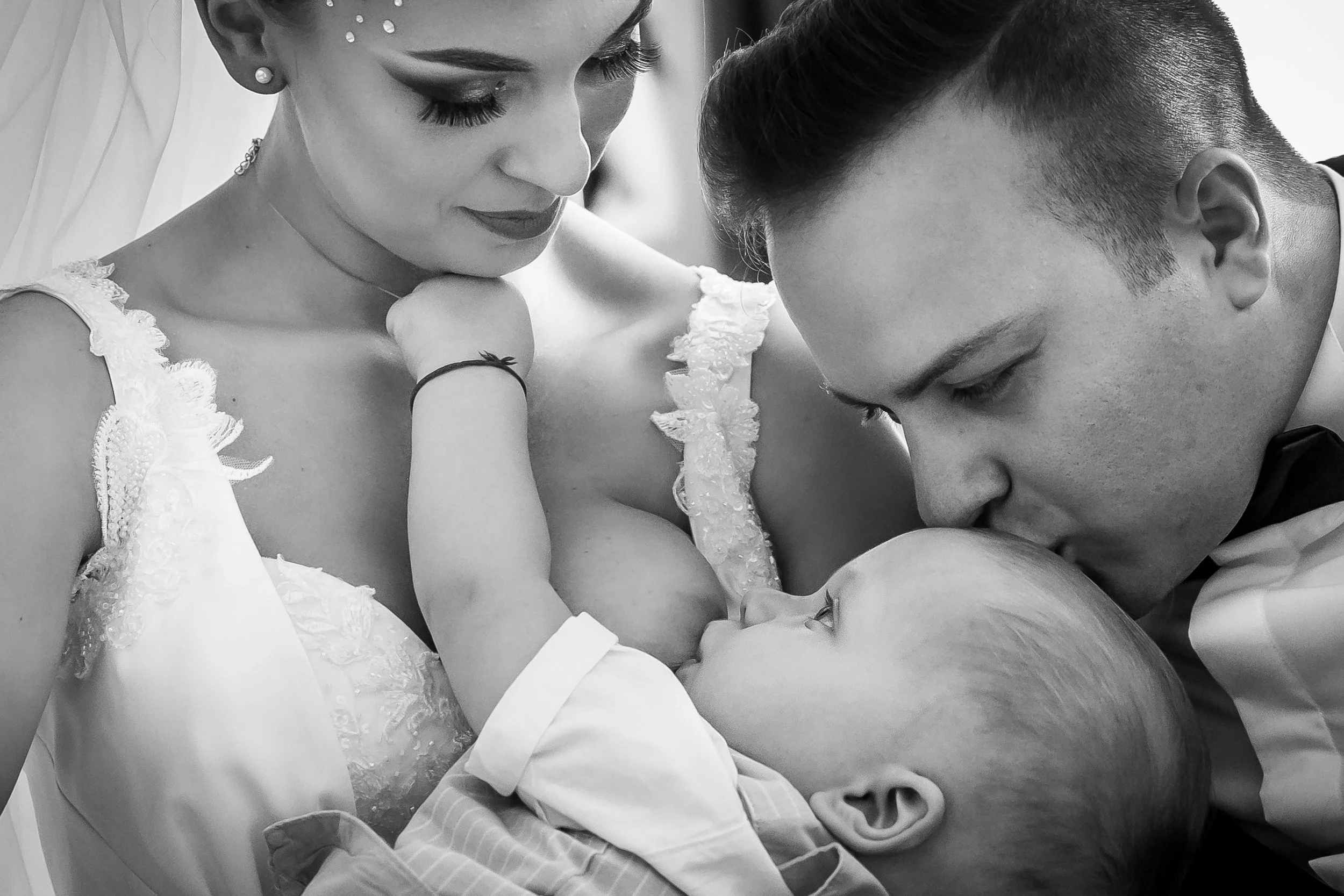 A black and white photo of a bride, groom, and a child sharing an intimate moment at a wedding. The bride is wearing a lace dress with floral details, and jewelry including earrings and a necklace. The groom is gently touching the child's face, while