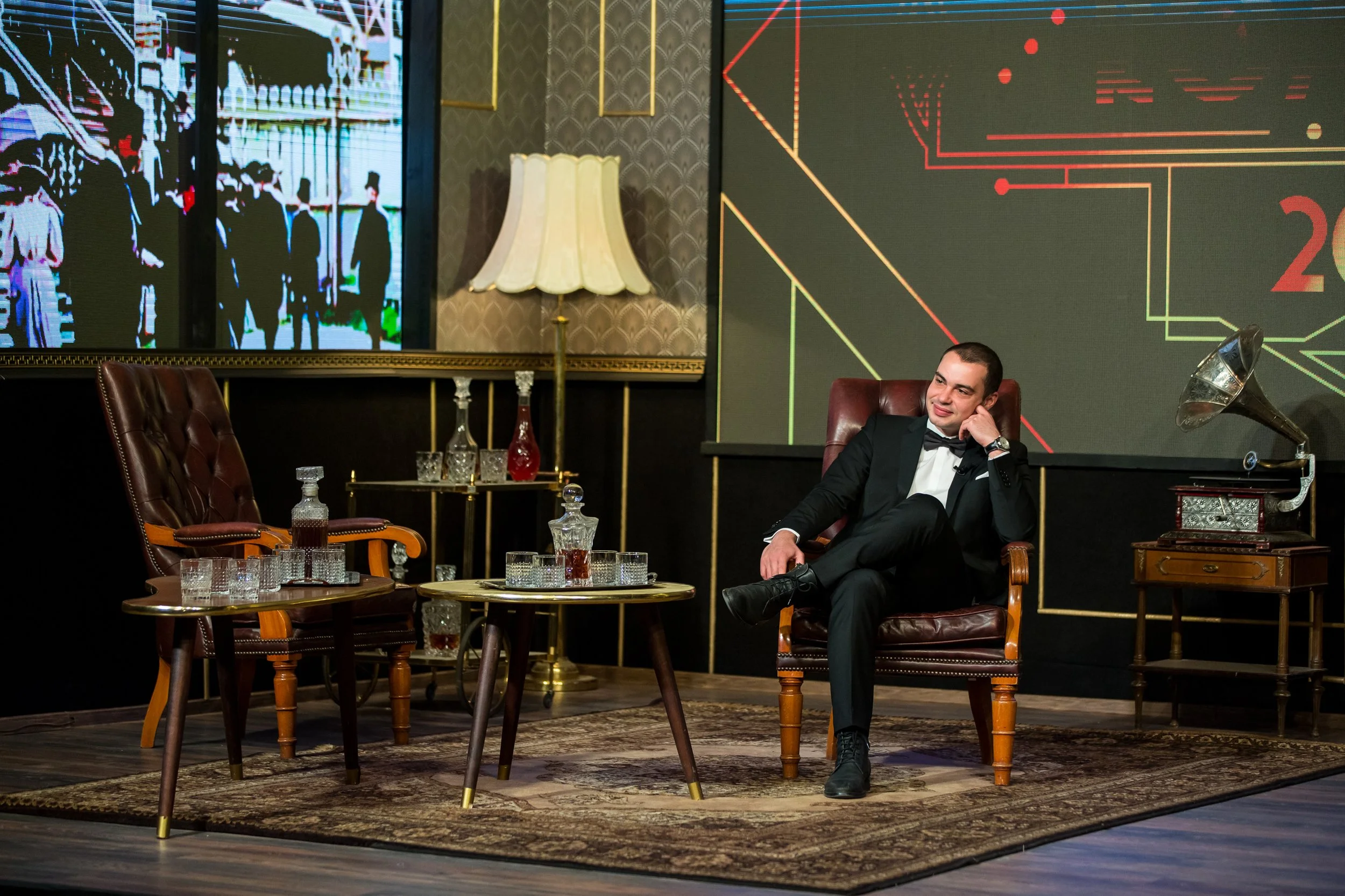 A man in a black tuxedo sitting in a vintage leather armchair in a well-decorated room. The room features a large digital screen displaying graphs and images, a vintage gramophone, a gold-colored lamp, and glassware on tables. The setting has a sophi