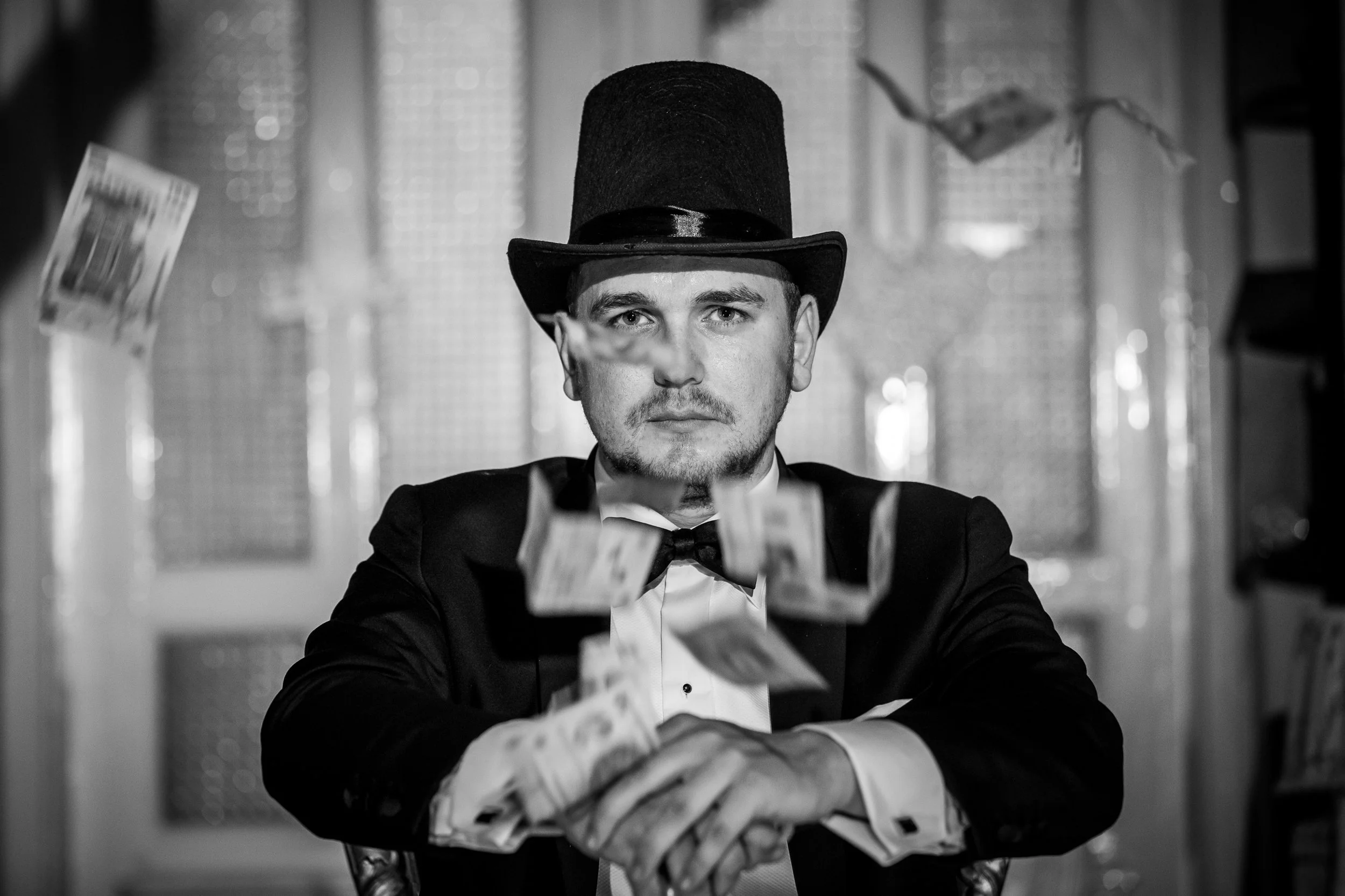A man dressed in formal attire, wearing a top hat, is sitting at a table and counting money with bills laid out in front of him. The setting appears to be luxurious and elegant.