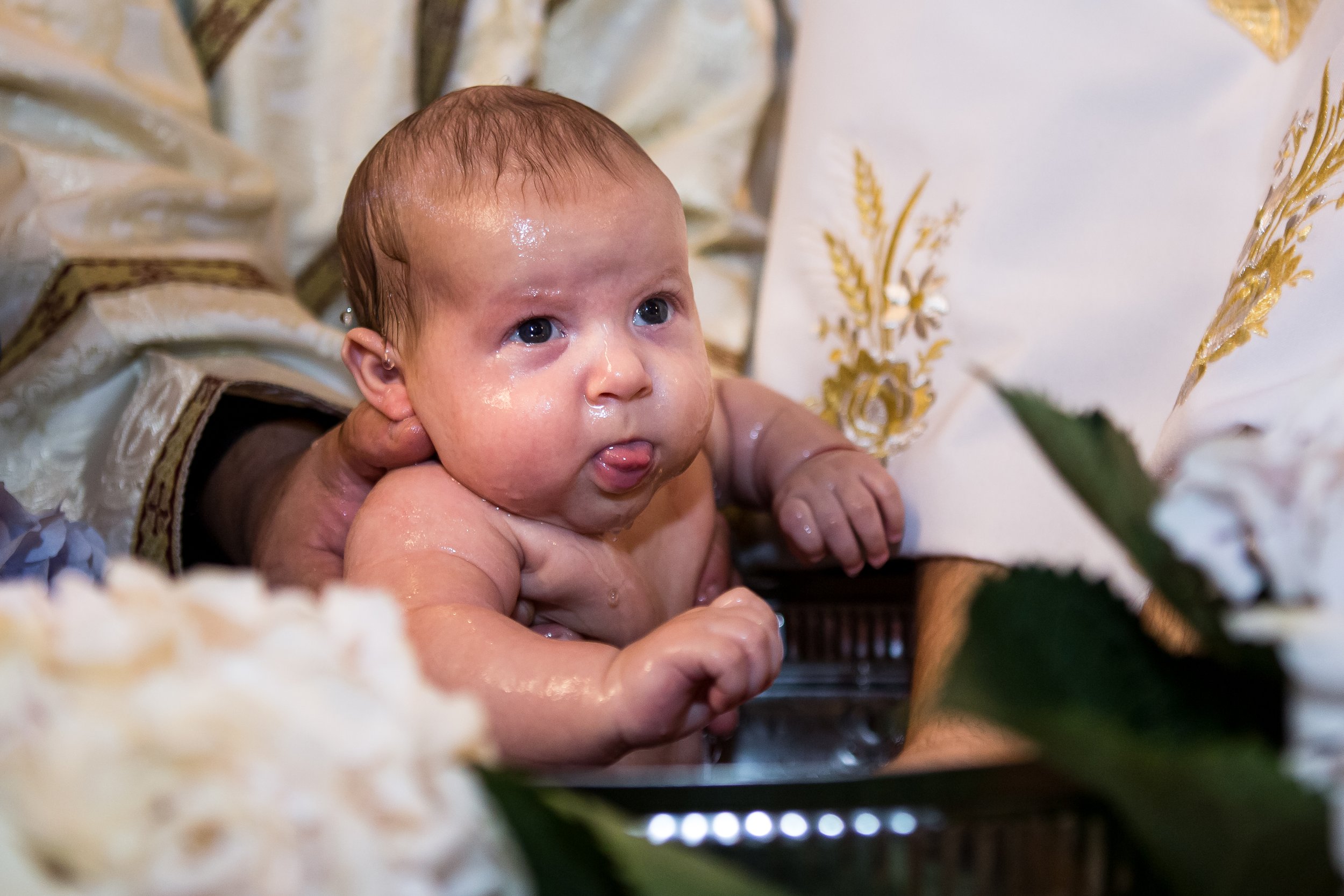 A baby with wet hair and blue eyes during a baptism or religious ceremony, being held by an adult, surrounded by flowers and ornate fabric.