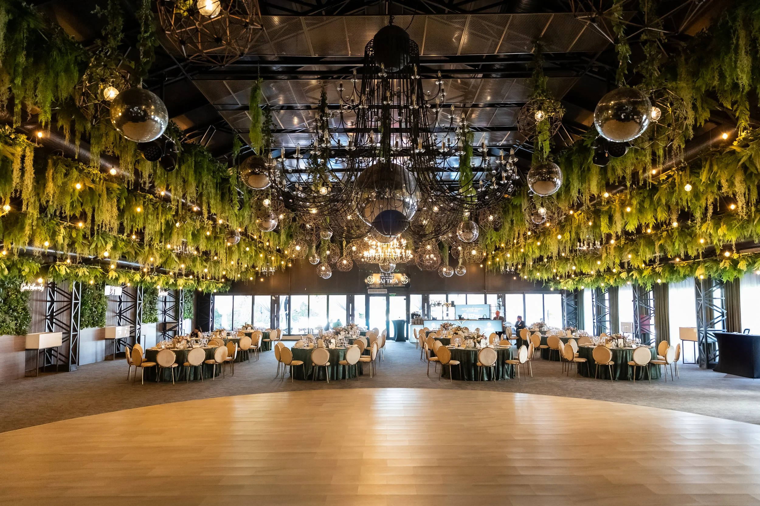 A banquet hall decorated with hanging greenery, chandeliers, and disco balls, set up for an event with round tables and beige chairs, near large windows.