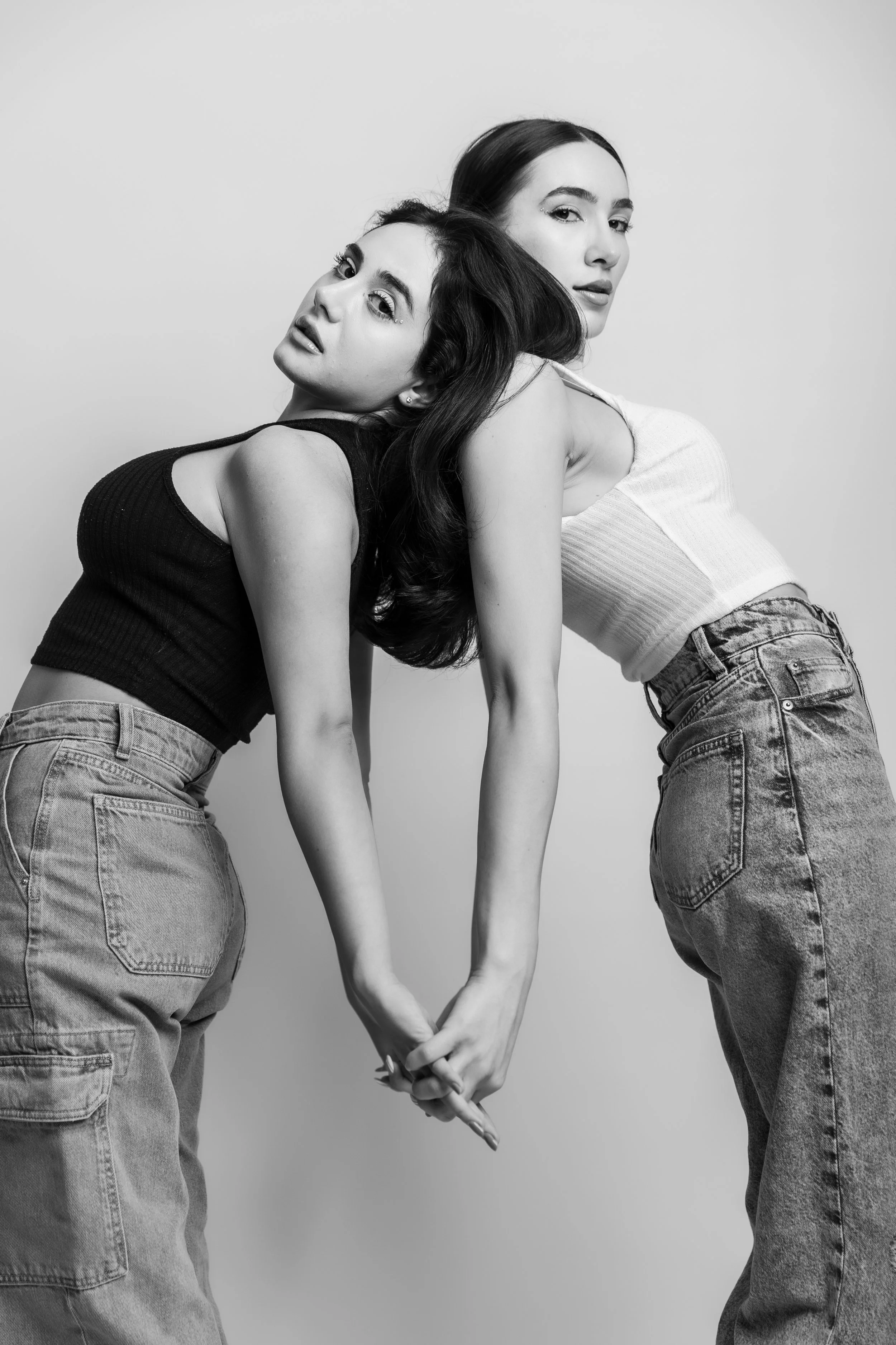 Black and white photo of two women with long dark hair, wearing casual denim jeans and sleeveless tops, holding hands and leaning against each other, looking at the camera.