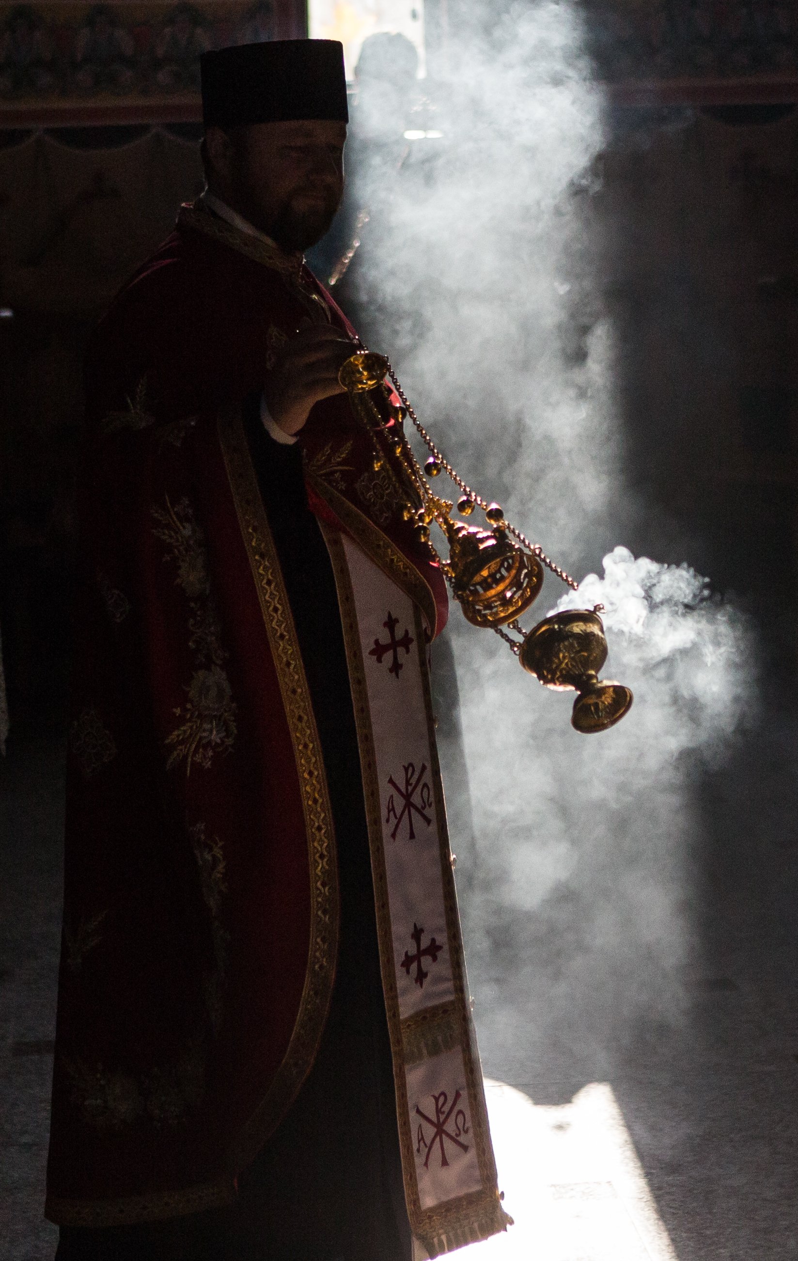 A man dressed in ornate religious robes holding a censer, with smoke rising around him, likely during a religious ceremony or ritual inside a church.