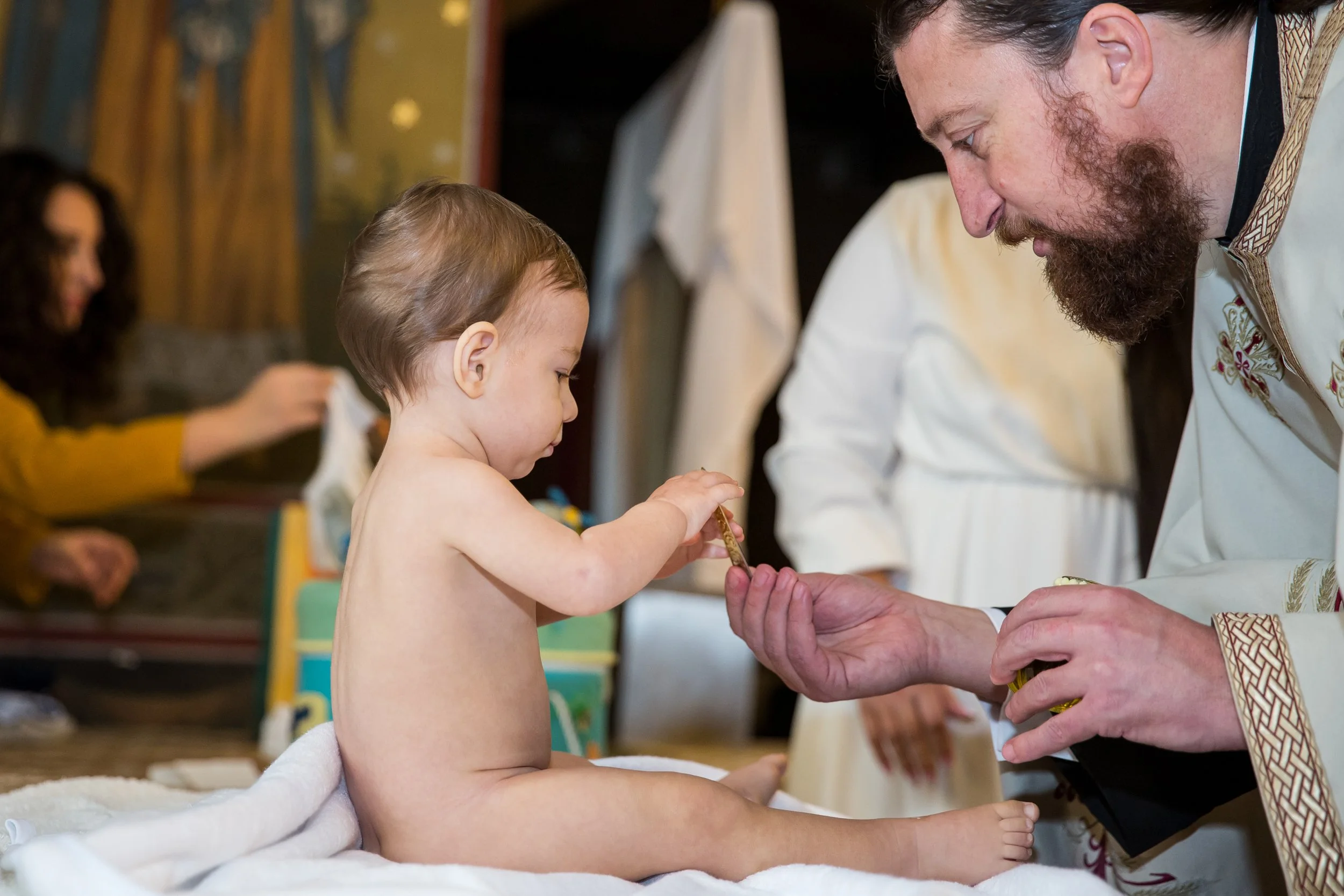 A young boy sitting shirtless on a bed interacts with an older man wearing ceremonial robes, possibly during a traditional or cultural ceremony. A woman in a yellow sweater is visible in the background, focused on her phone.