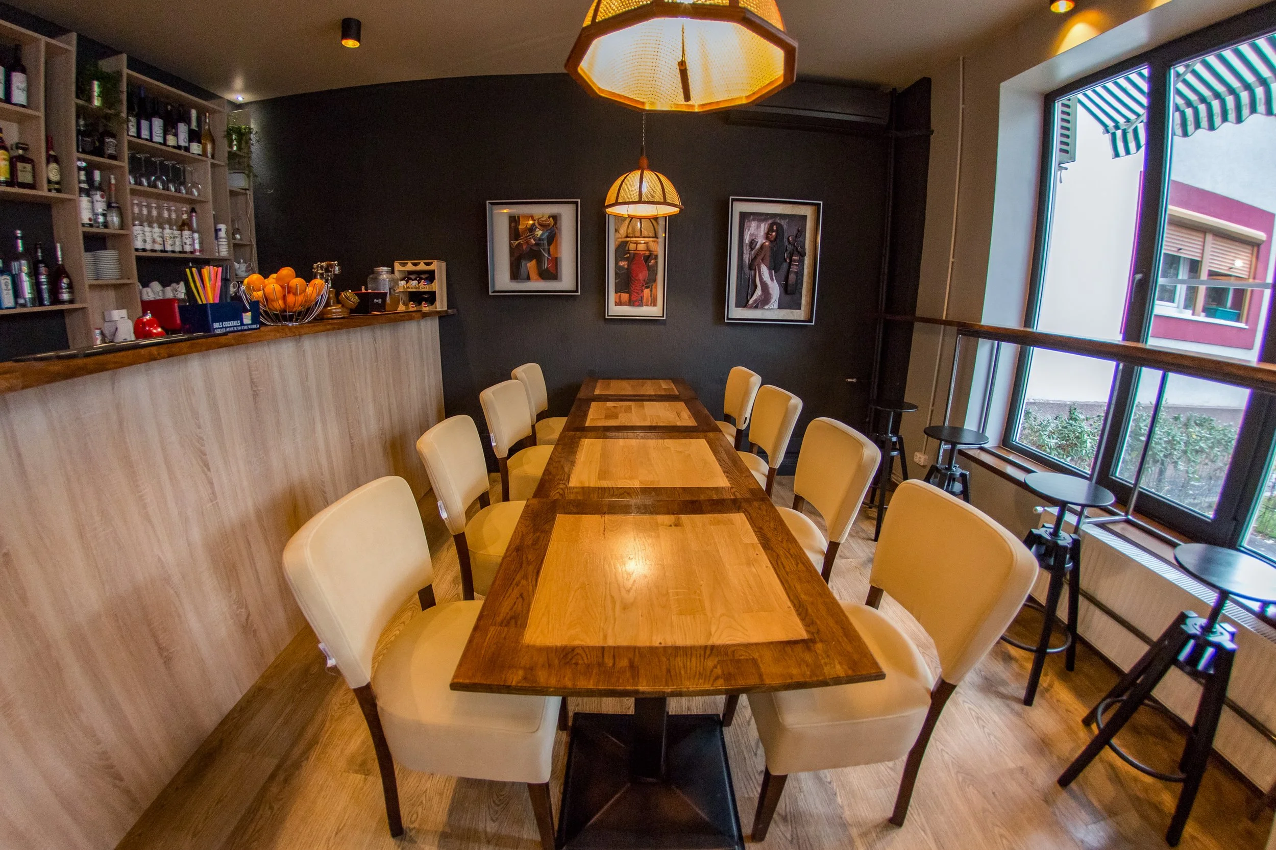Interior of a restaurant with a long wooden table and yellow upholstered chairs, dark accent wall with artwork, large windows with striped awnings, hanging pendant lights, bar area with bottles, glasses, and fruit bowls.