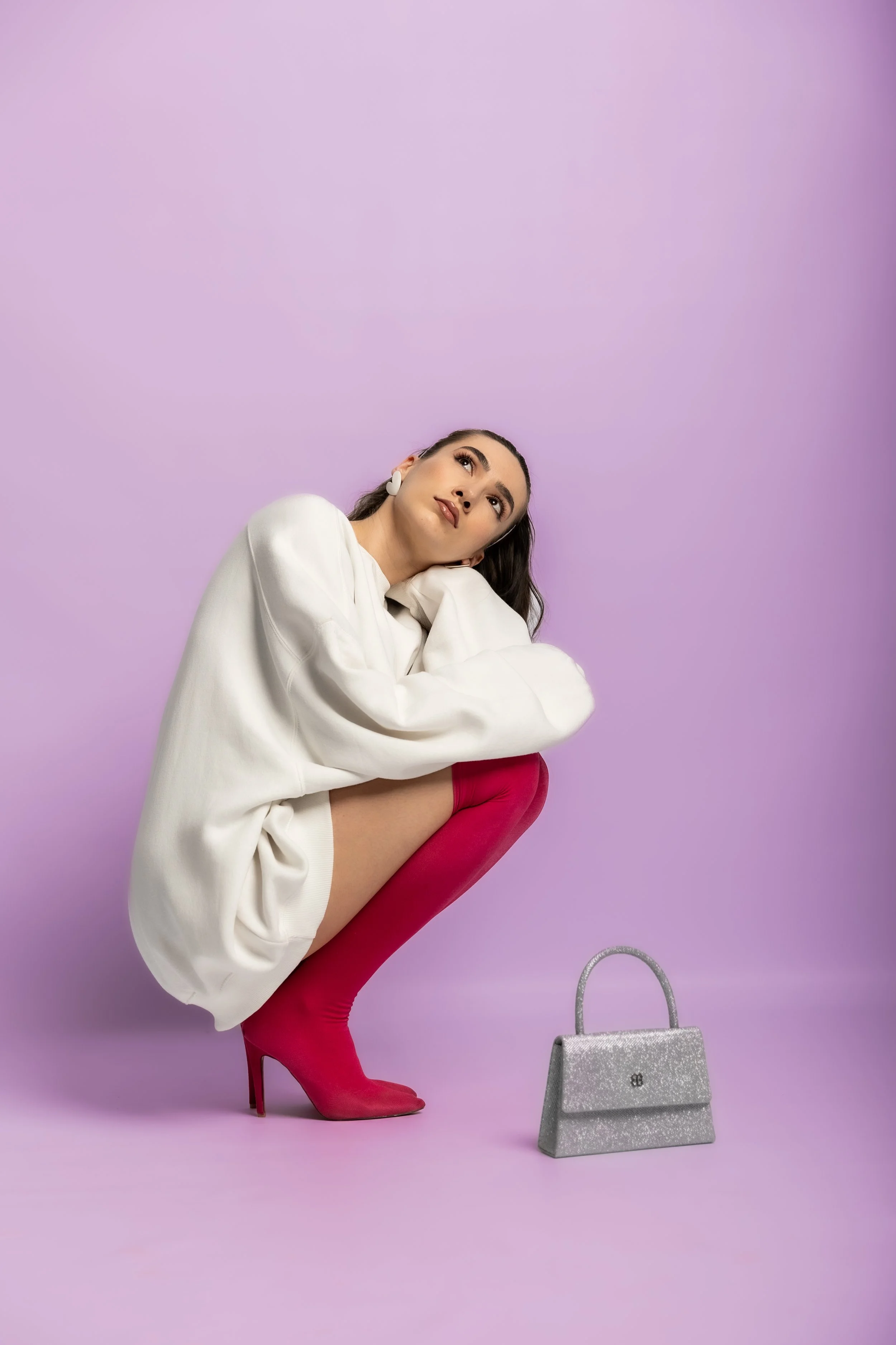 A woman with dark hair and makeup, sitting on a high pink stiletto heel, wearing a white oversized shirt, pink thigh-high boots, and large white earrings, against a lavender background, with a silver glitter handbag on the floor.