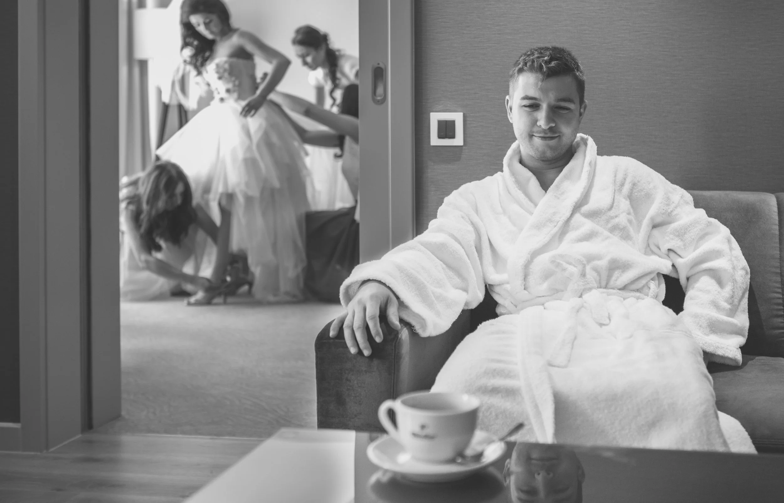 A man sitting on a chair in a bathrobe at a table with a cup and saucer, looking at a reflection of women preparing a wedding dress in a mirror. The women are in a room behind a sliding door.