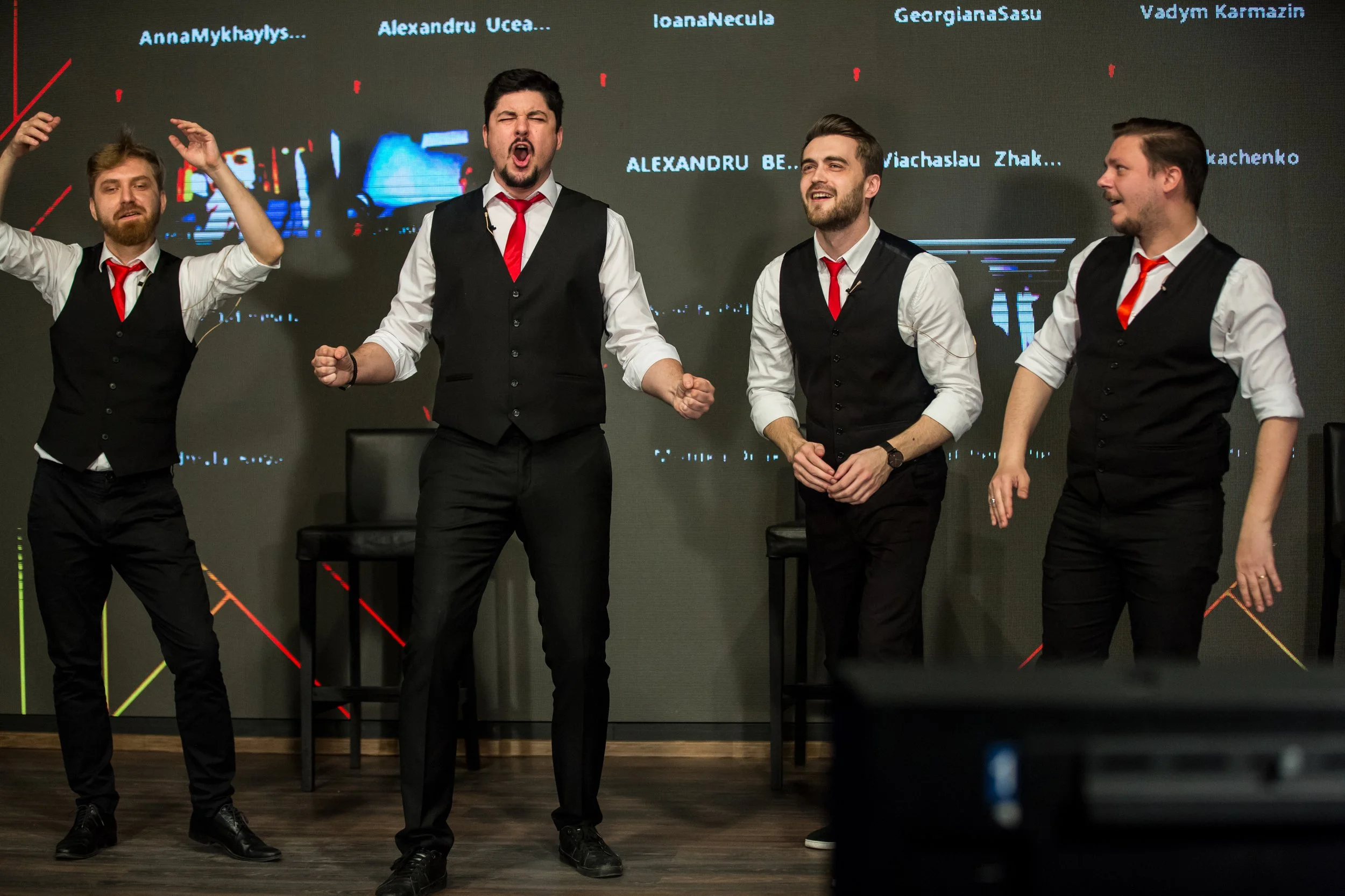 Four men with dark hair and beards, dressed in white shirts, black vests, and red ties, are standing on a stage with a digital screen behind them displaying names. They are singing or speaking passionately, with one man in the center raising his fist