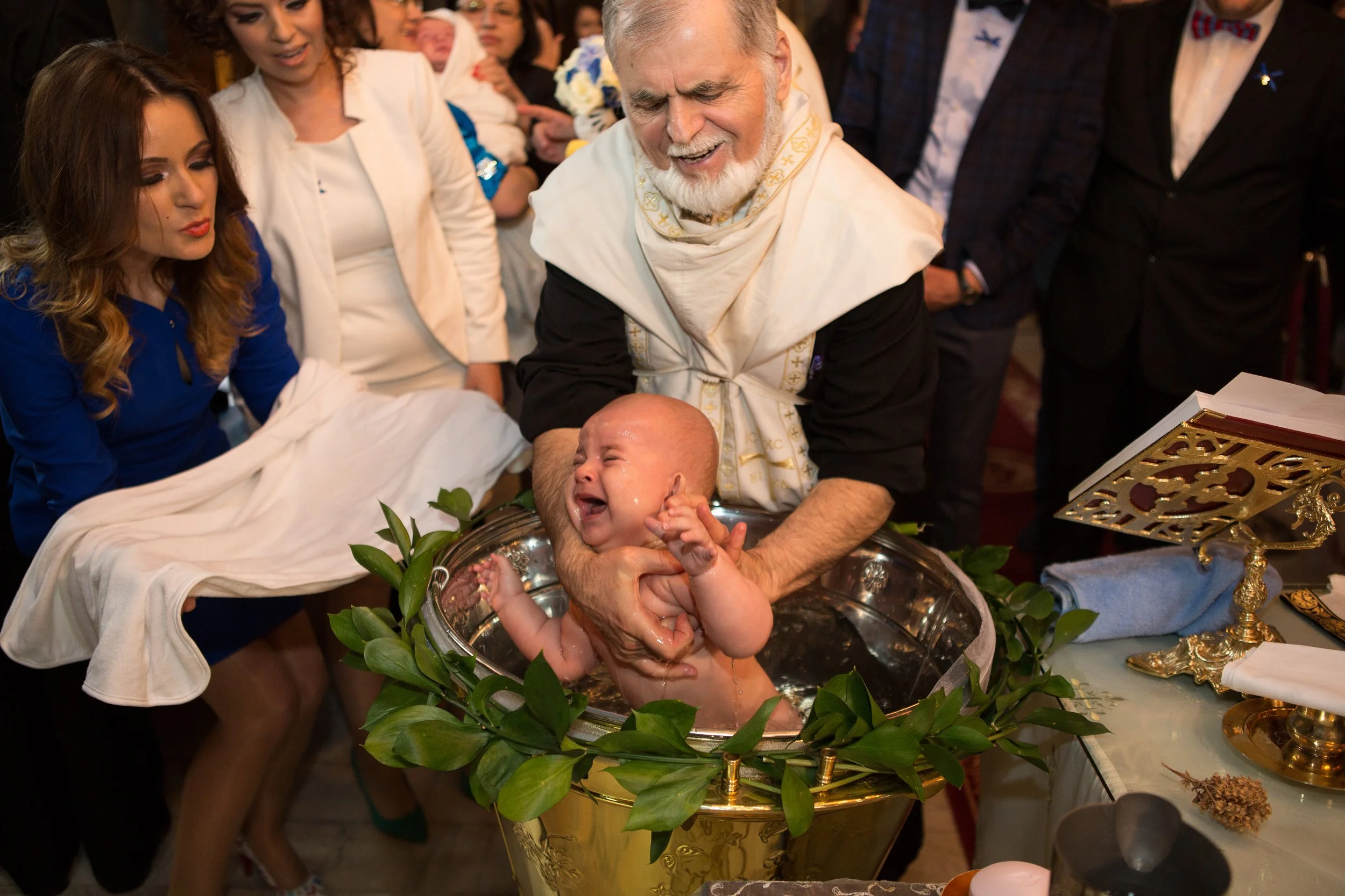 Baptism ceremony with a baby in a baptismal font held by an elderly man, surrounded by family and religious items.