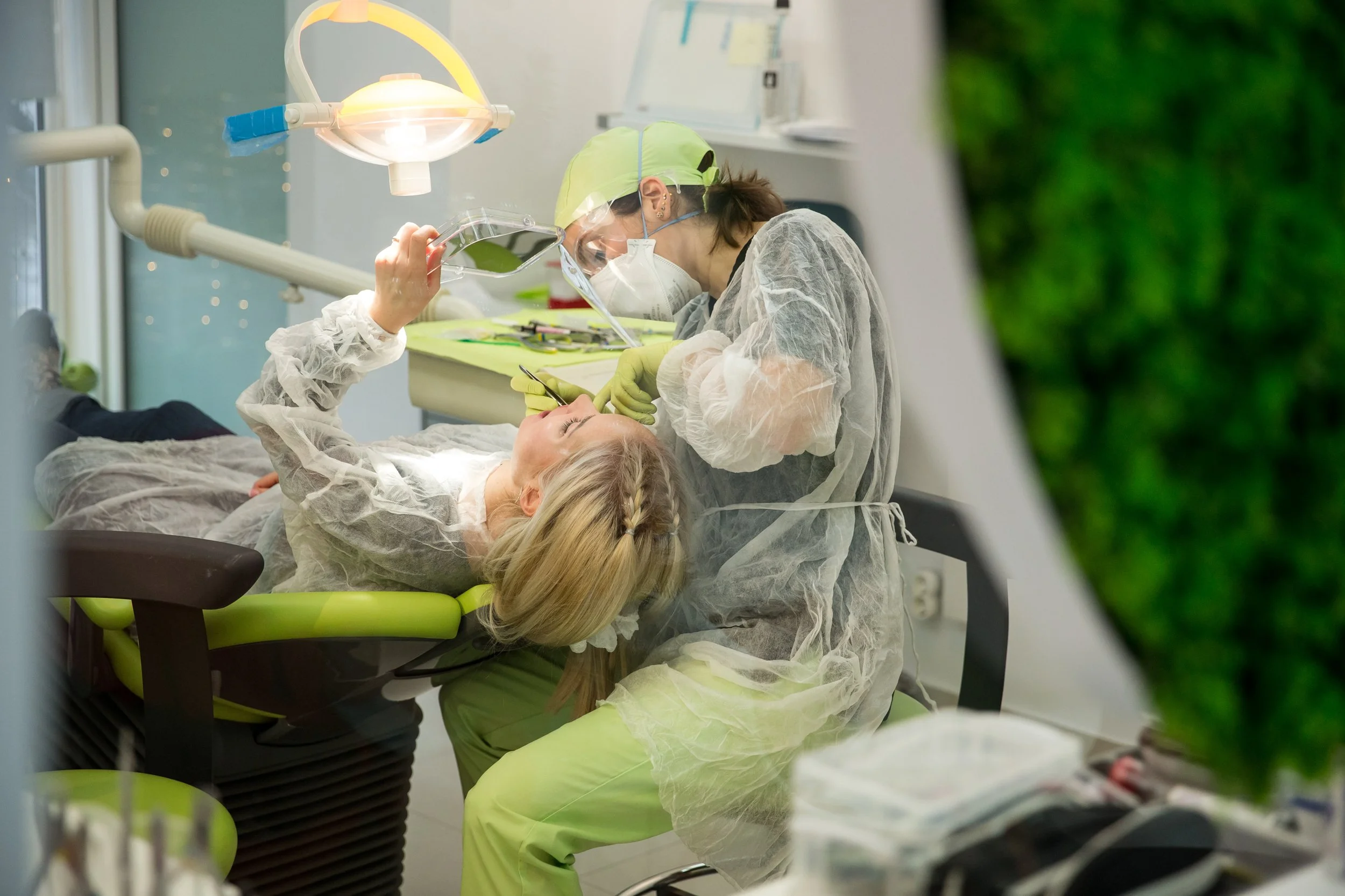 A healthcare worker wearing protective gear, including a mask, gown, gloves, and face shield, appears to be attending to a young woman lying in a hospital bed. The young woman has blonde hair, is wearing a gown, and is holding a phone while looking a