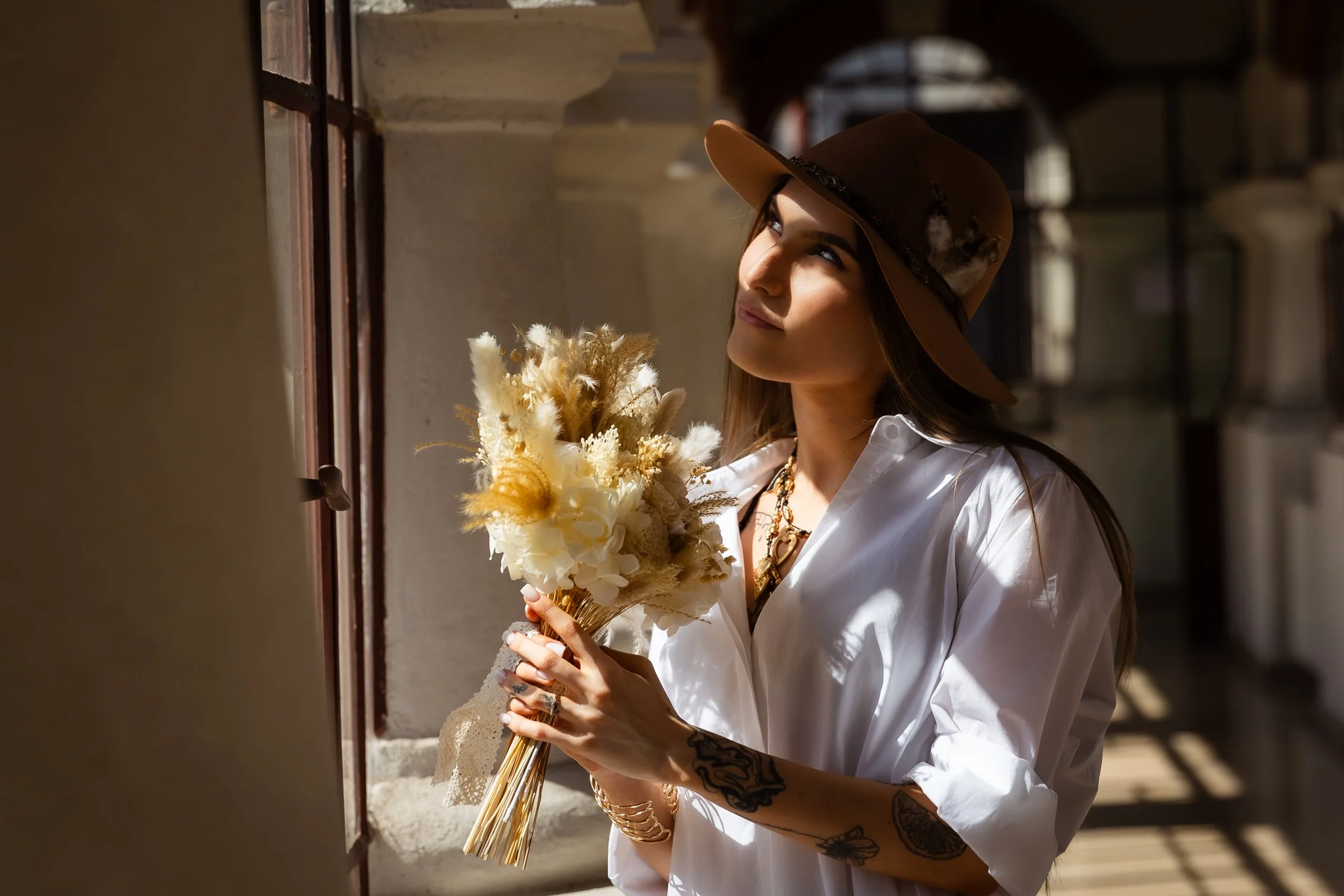 Woman with tattoos on her arms, wearing a wide-brimmed hat and a white shirt, holding a bouquet of dried flowers, looking out a window with sunlight streaming in.