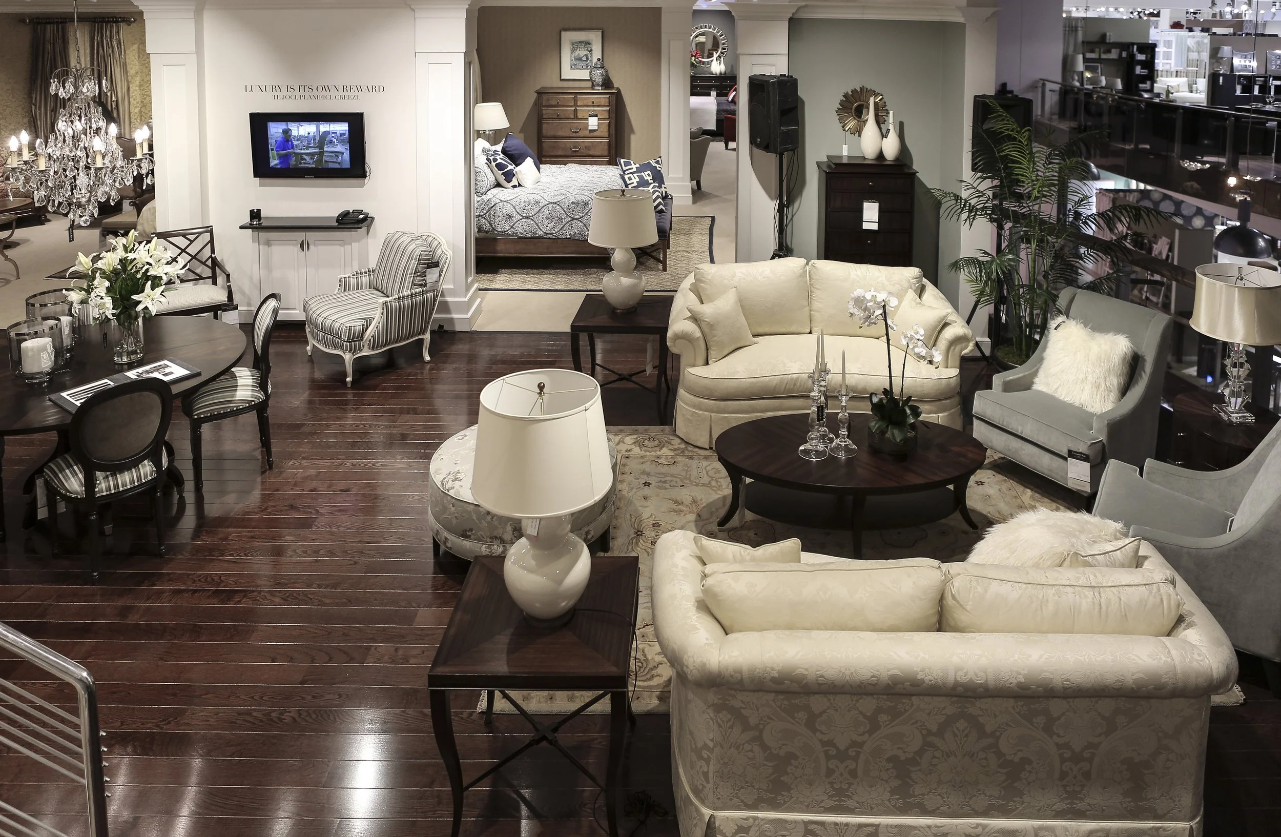 A furniture showroom displaying various living room and bedroom furniture including sofas, chairs, tables, and lamps. The room has dark wooden floors, with some furniture decorated with flowers and candles, and a wall-mounted TV in the center.