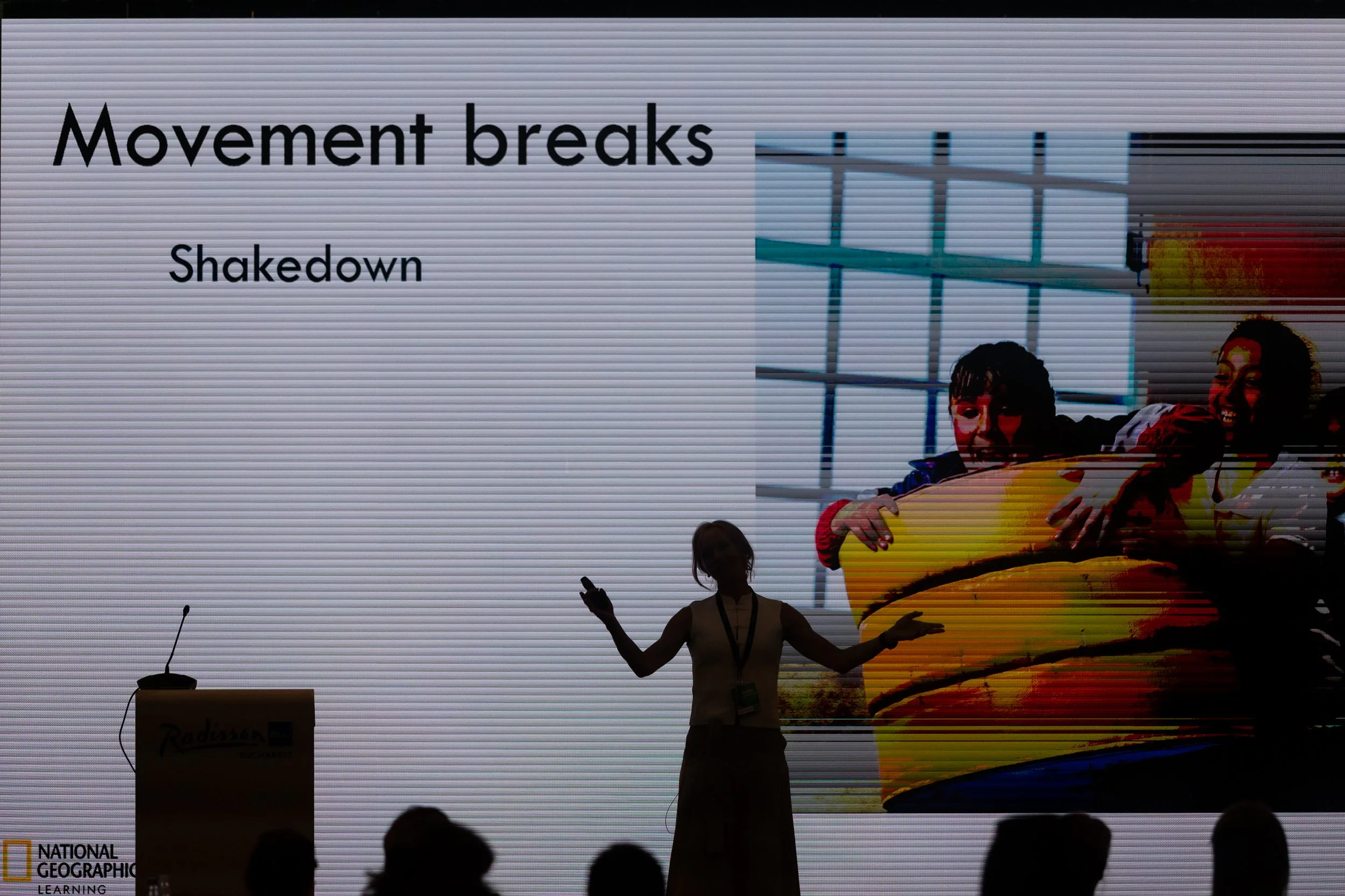 A woman stands on stage in front of a large screen displaying the text 'Movement breaks' and 'Shakedown.' She is gesturing with her arms, with a microphone and a hand-held device. The stage has a podium with the 'National Geographic Learning' logo an