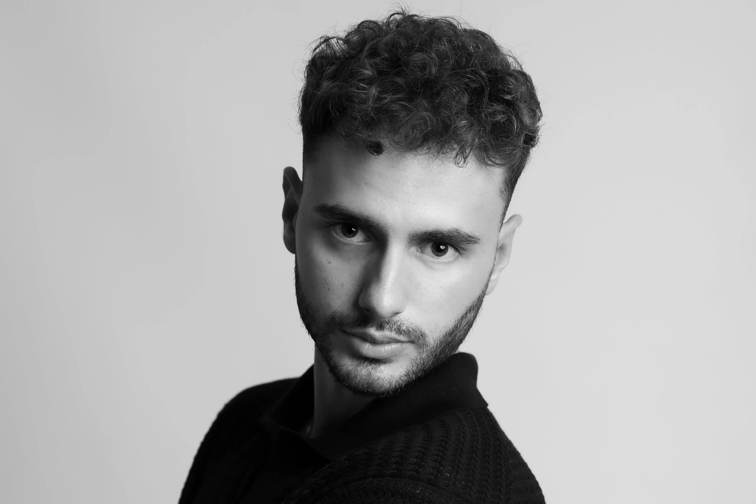 Black and white portrait of a young man with curly hair and a beard, looking into the camera against a plain background.
