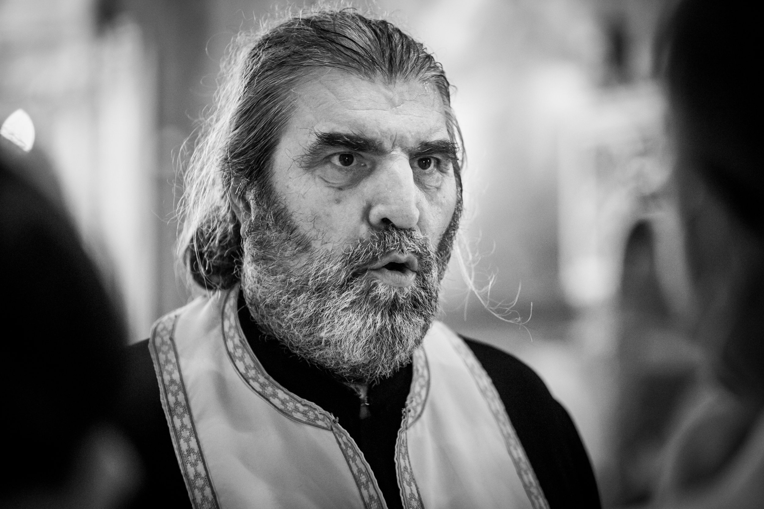 A black and white close-up photograph of a bearded man with long hair, wearing a traditional embroidered garment, engaged in conversation or thought.