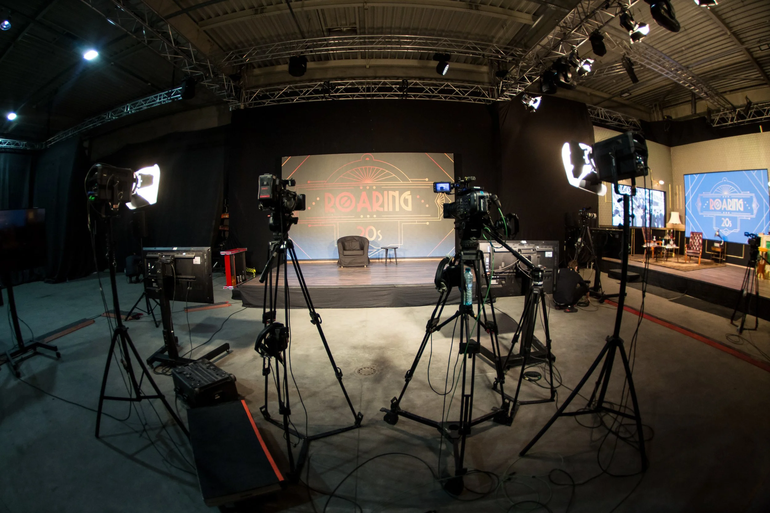A stage setup with multiple cameras and lighting equipment, a large screen displaying the words 'ROARING 20s', and chairs arranged for a presentation or event in an indoor venue.