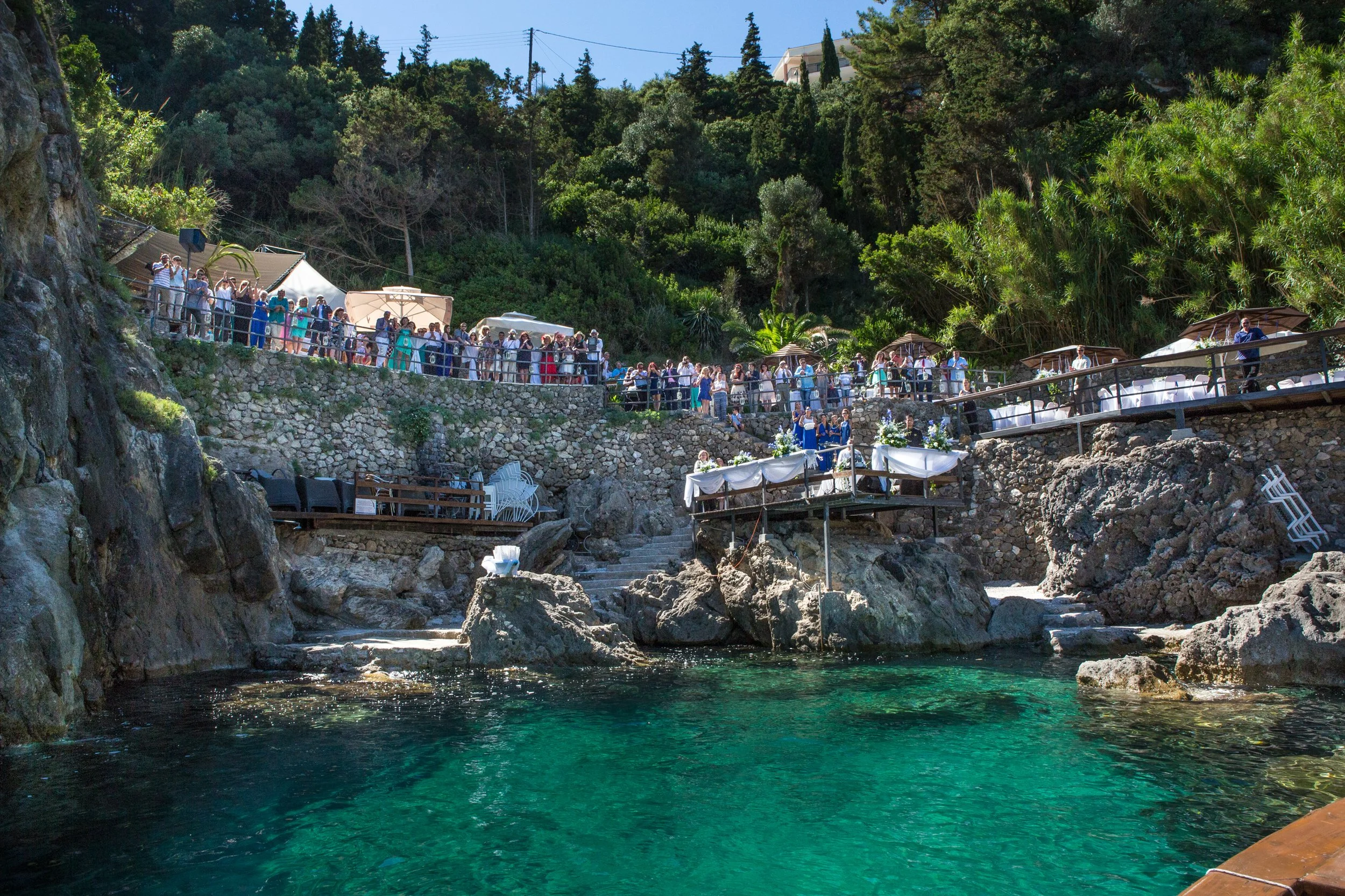 A group of people at an outdoor event along a rocky coastline, with tables, umbrellas, and lush green trees in the background on a sunny day.