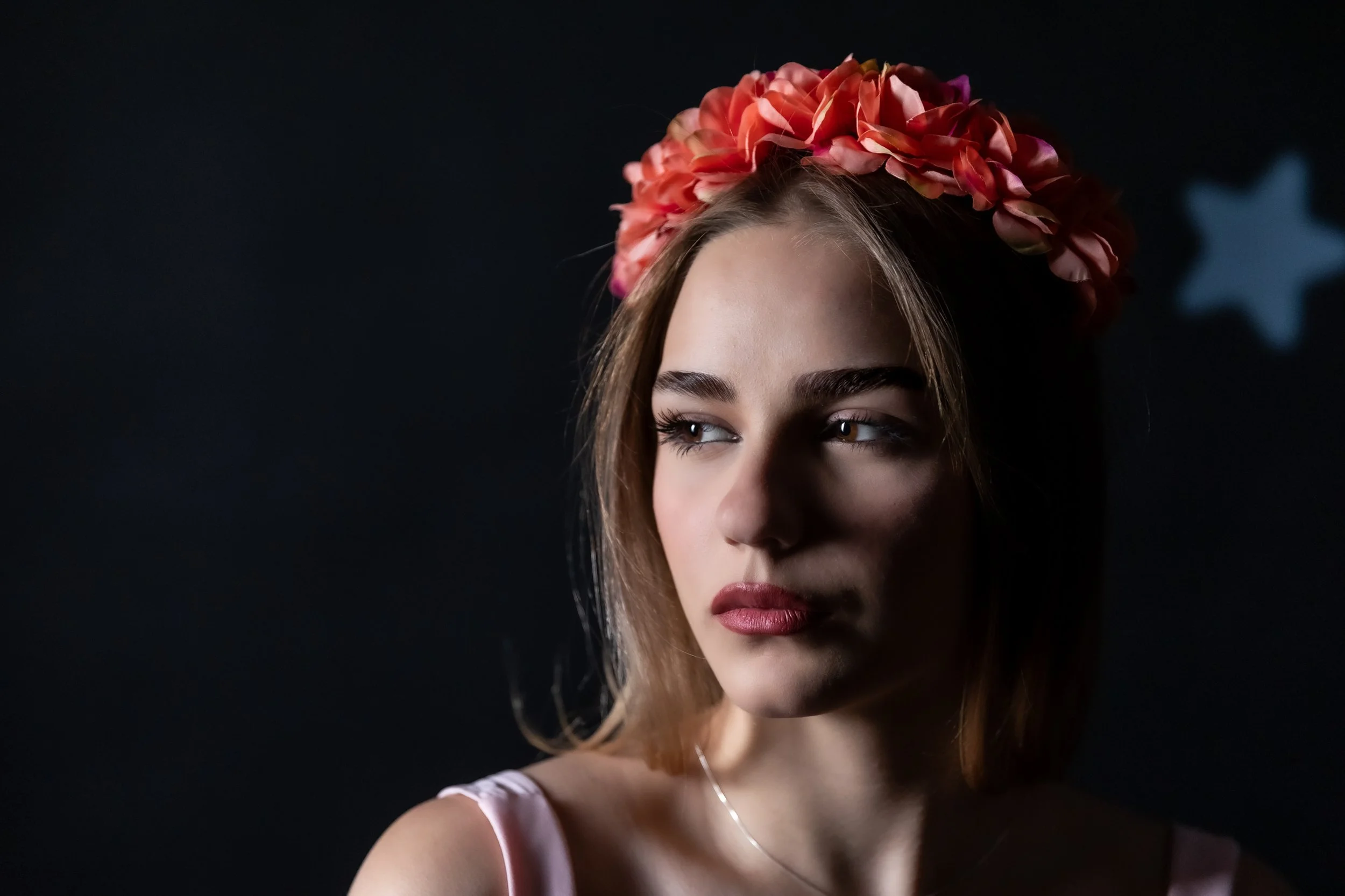 A young woman with light brown hair, wearing a pink satin top, a floral crown of pink and orange petals, and a delicate necklace, posing against a dark background with a blurred star-shaped decoration.
