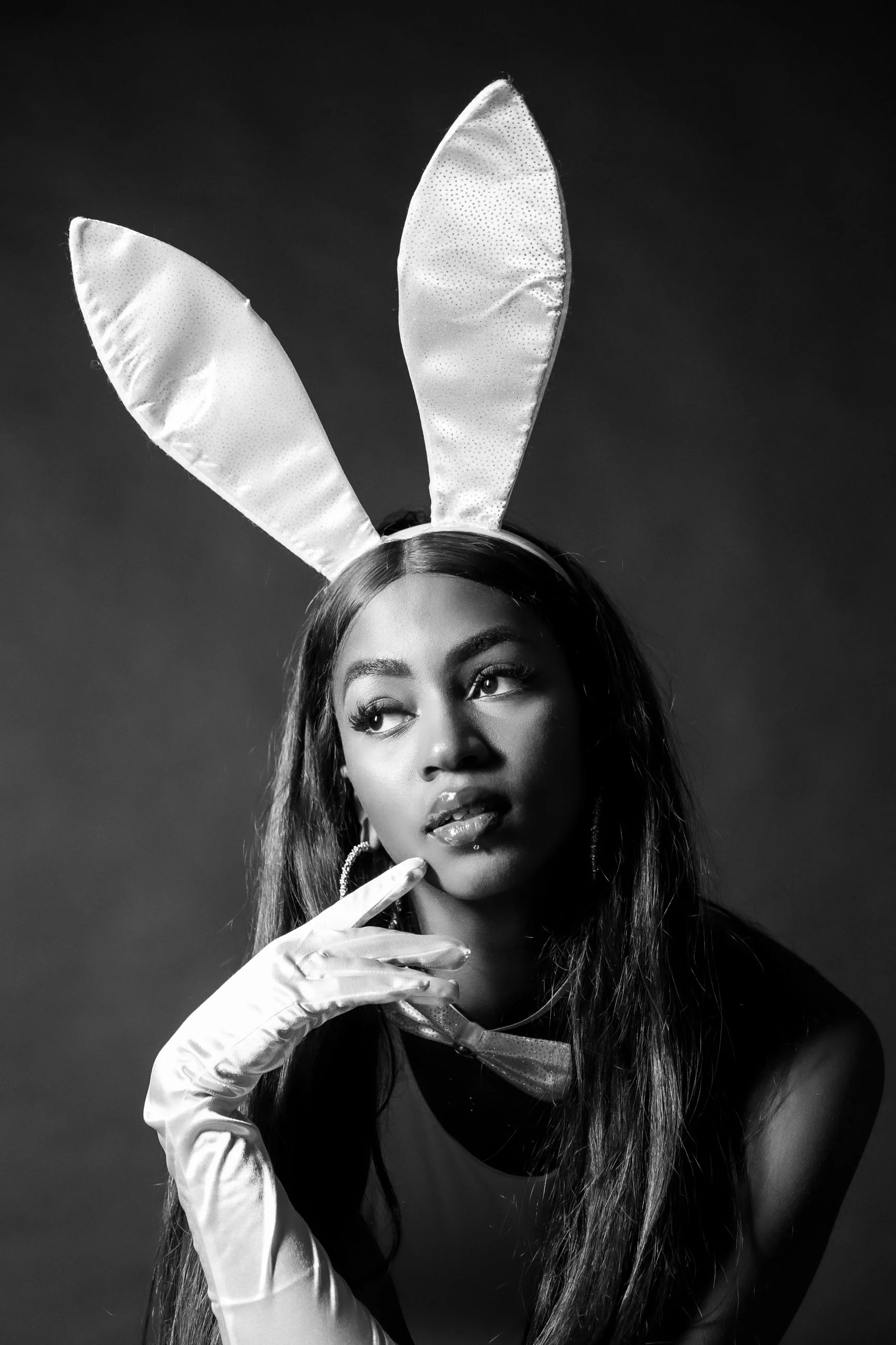 A woman with long hair and hoop earrings wears bunny ears, satin gloves, and a dark top, looking thoughtful with her fingers near her face against a dark background.