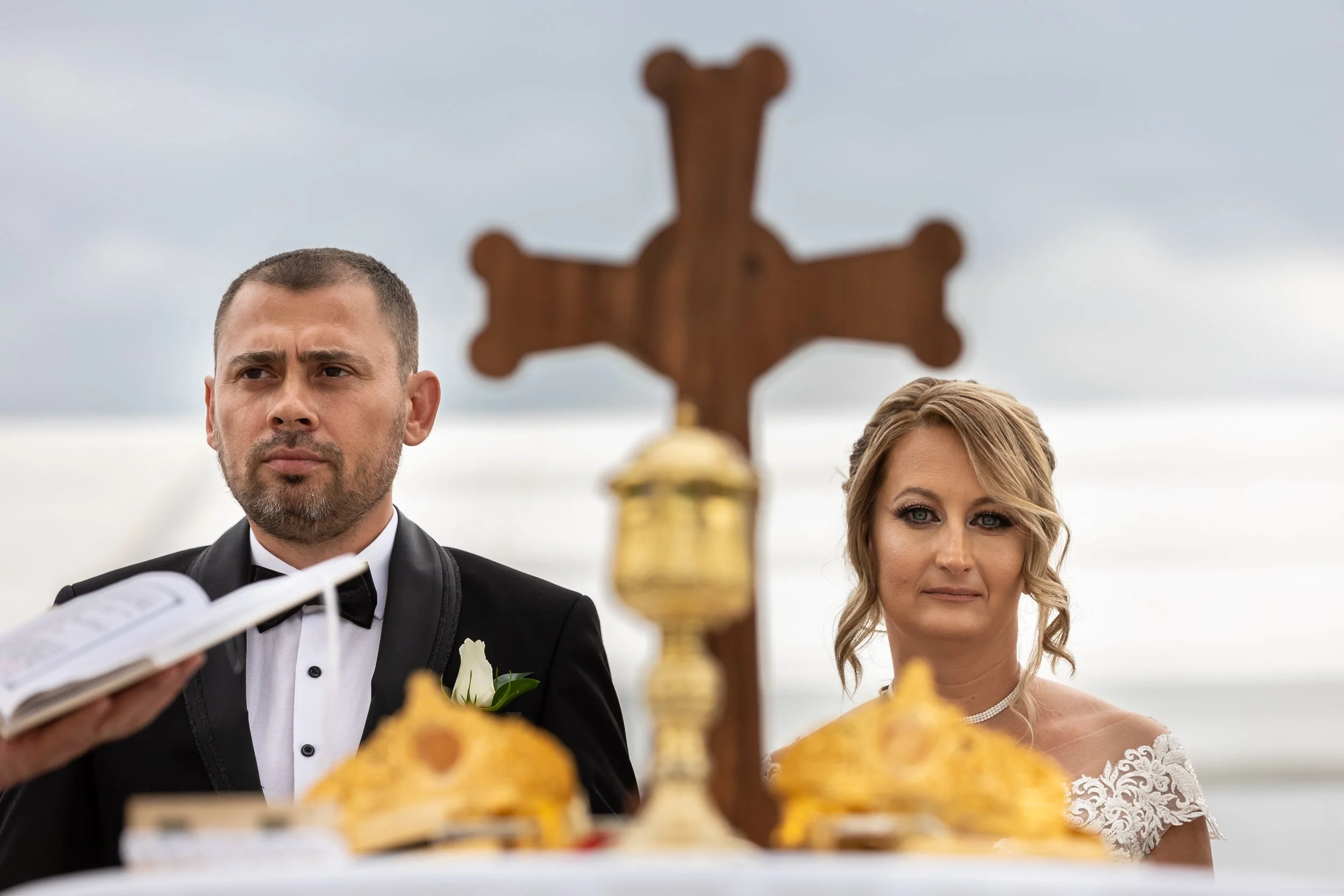A man in a tuxedo and a woman in a wedding dress standing outdoors near a cross-like structure, with a cloudy sky in the background.