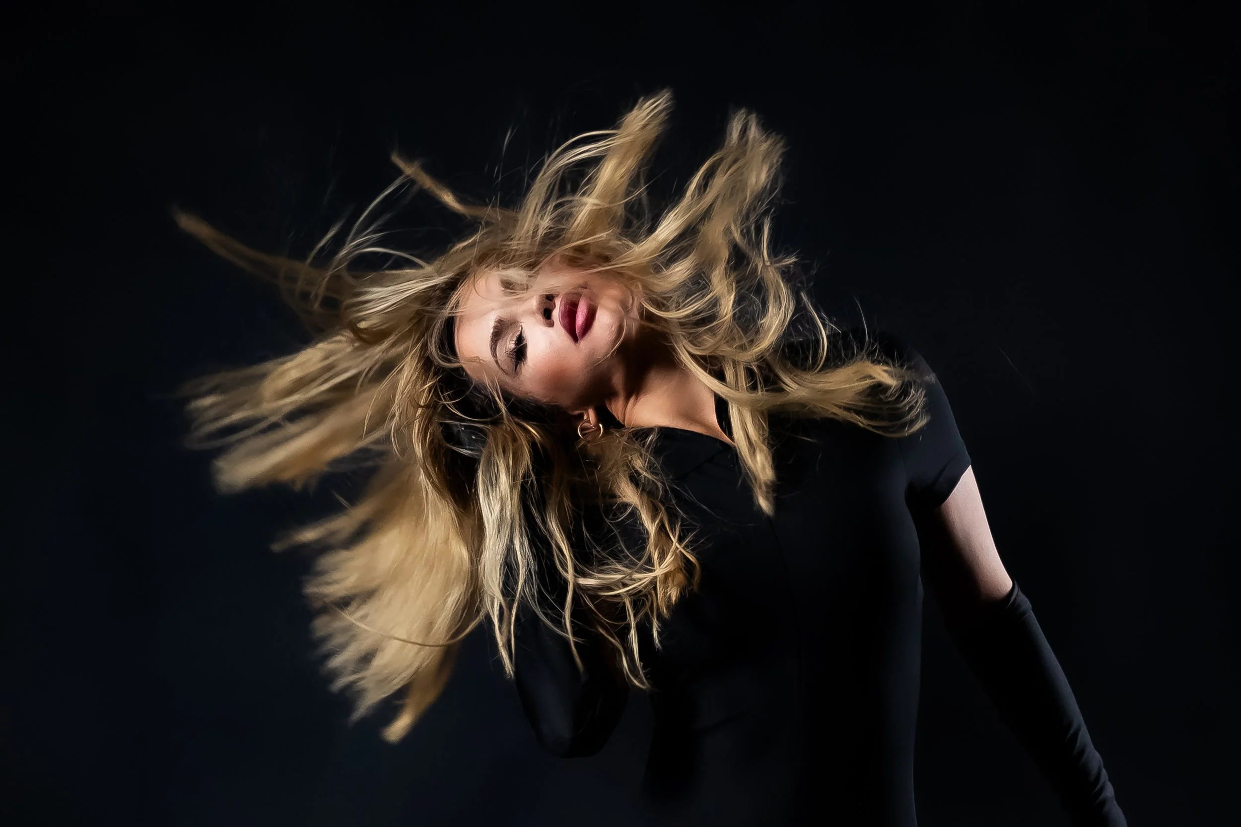 A woman with long blonde hair, wearing a black outfit, leaning to the side with her head tilted back against a black background.