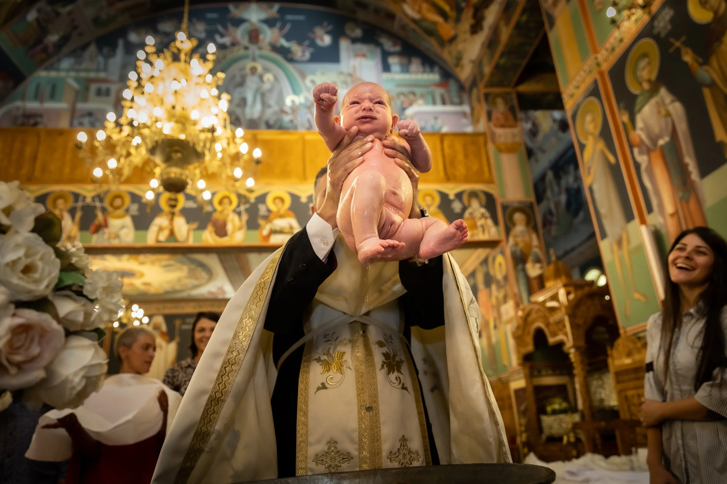 A baby being baptized in an ornate church, raised by a priest in religious vestments during a ceremony, with people watching and a chandelier overhead.