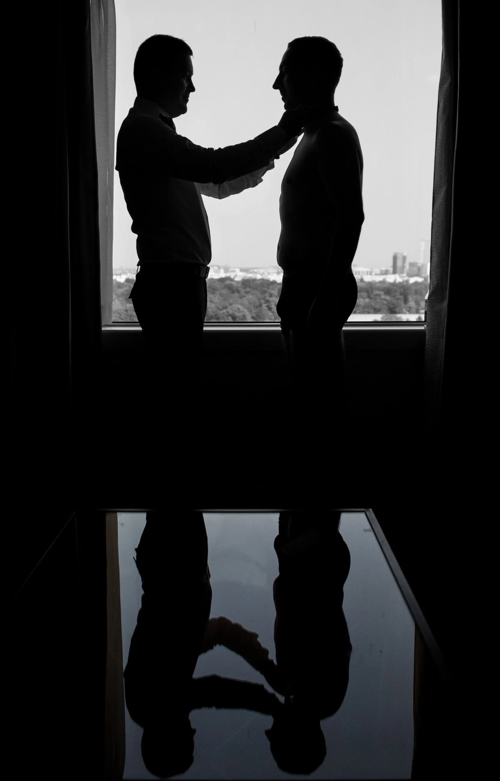 Two men in suits are facing each other near a large window with a cityscape in the background. One man appears to be adjusting the neck of the other, and both are silhouetted against the bright outside light, creating a mirror image on a reflective s