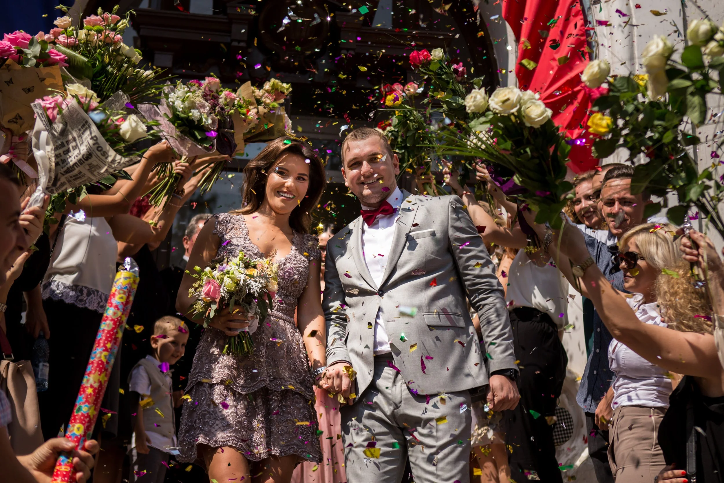A bride and groom walking hand-in-hand through a crowd during their wedding celebration, surrounded by guests and colorful confetti.