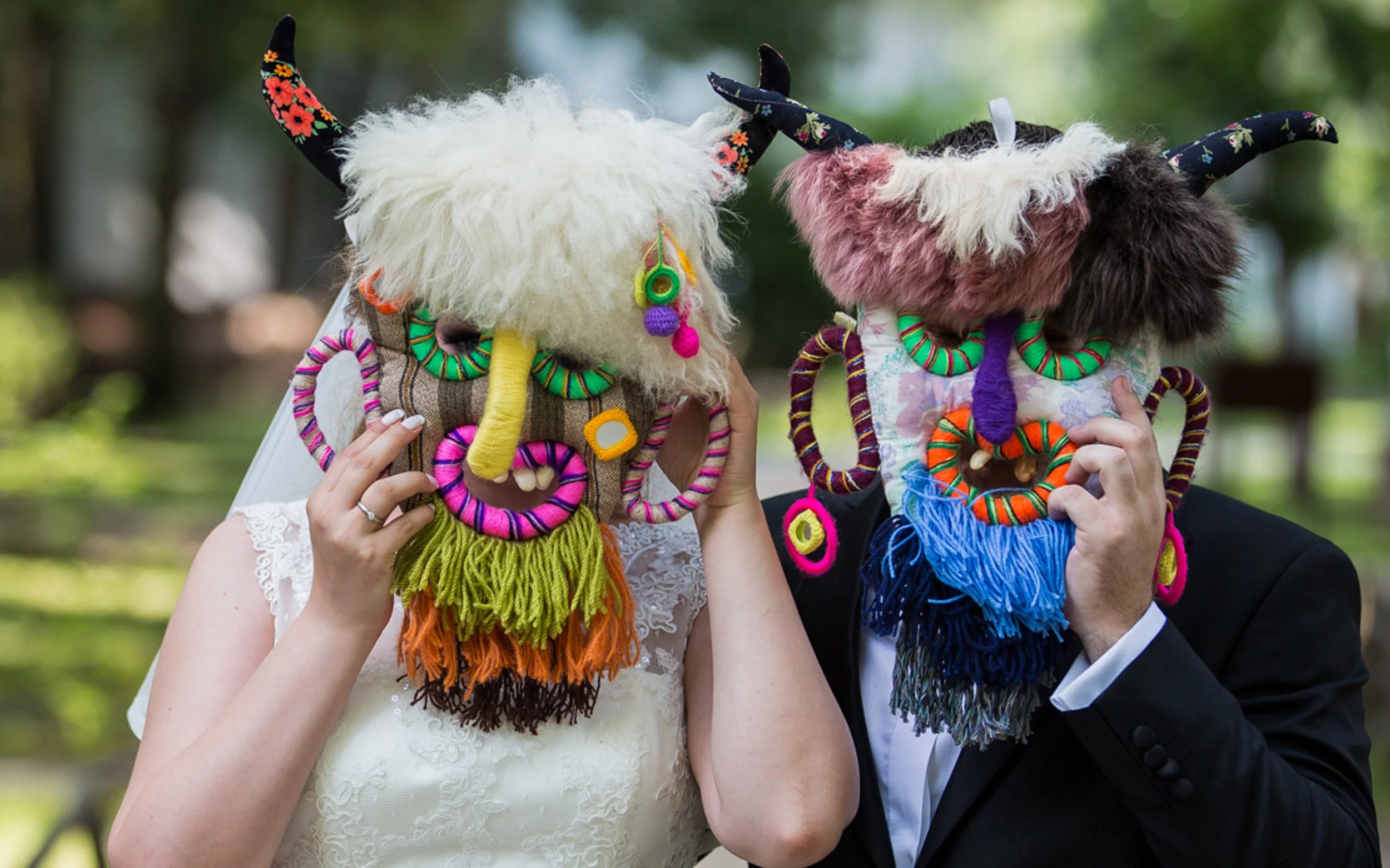 Two people holding colorful, furry masks with horns and yarn decorations over their faces, outdoors with a blurred green background.