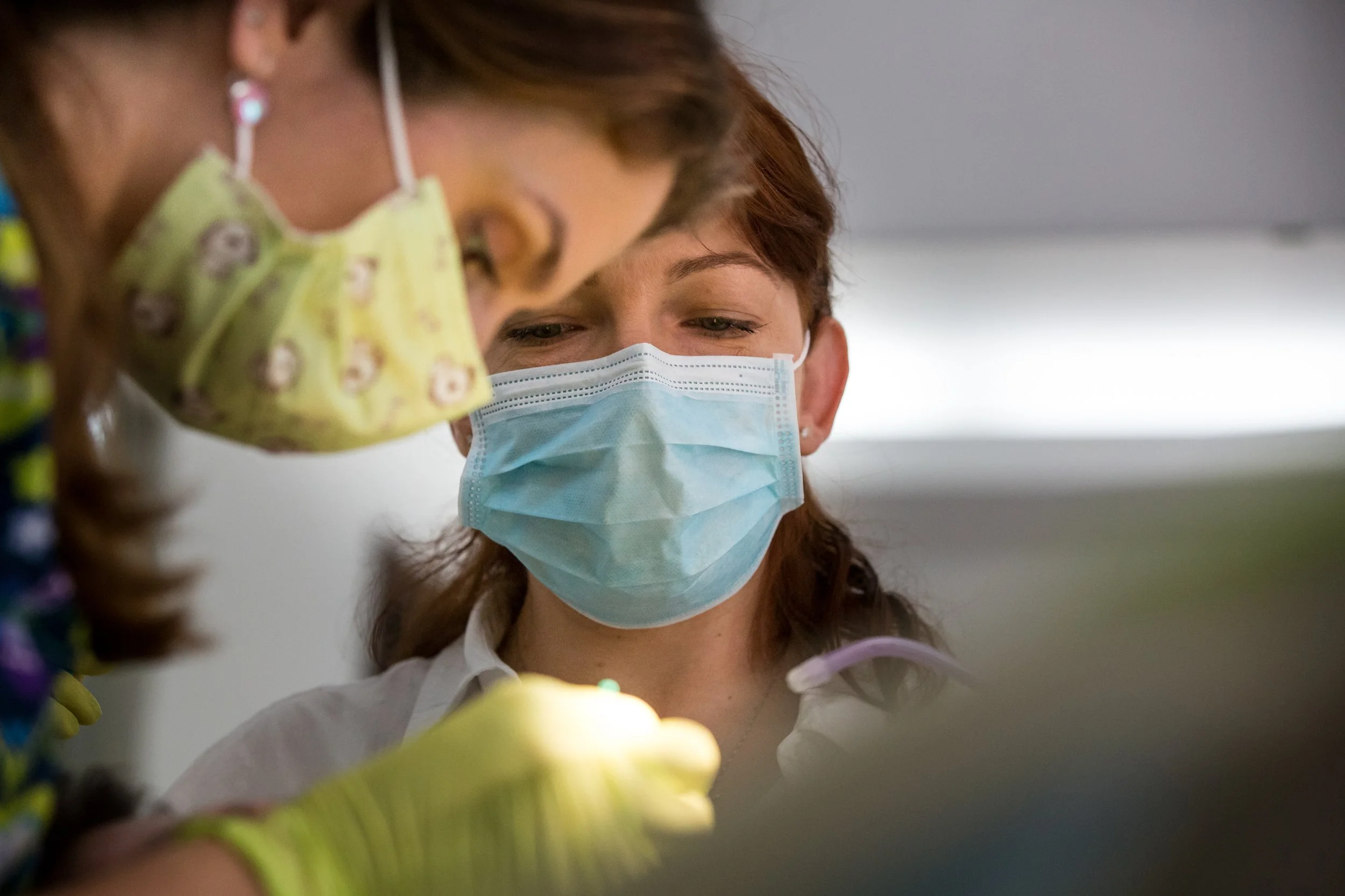 A woman with red hair wearing a blue surgical mask and white shirt interacting with a young girl. The girl is wearing a yellow mask with cartoon characters and colorful clothing, and is also engaging with the woman. The image depicts a close-up of th