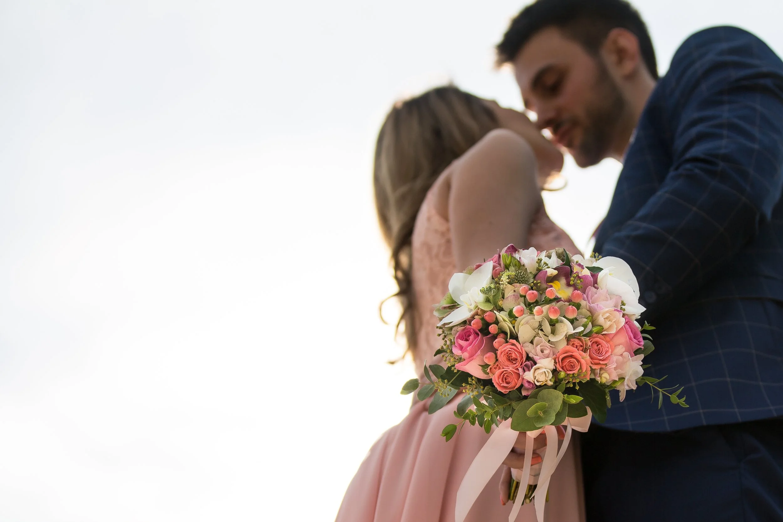 A couple embraces while holding a colorful bouquet of flowers, with a clear sky in the background.