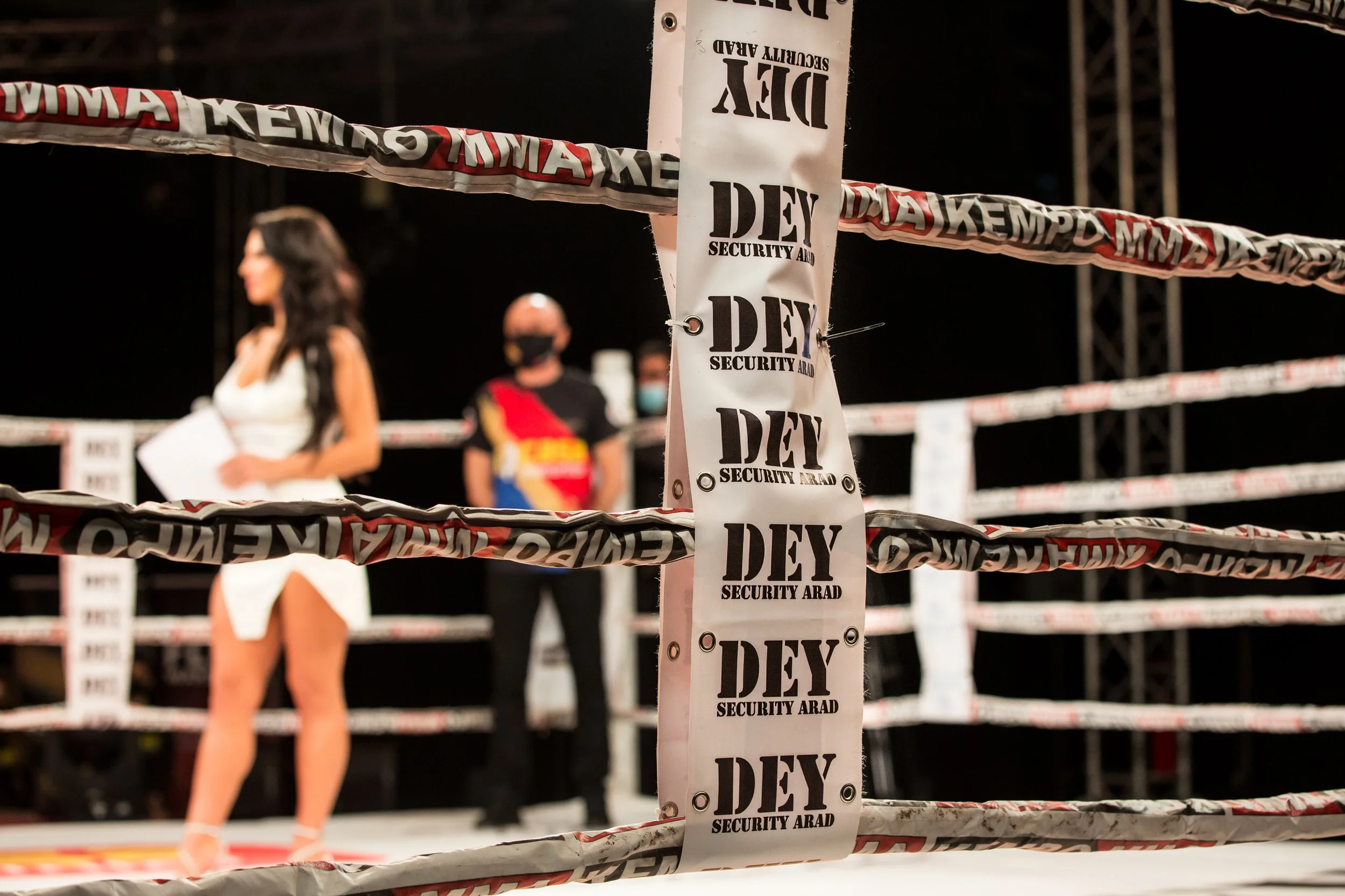 A boxing ring with security tape in the foreground that reads 'Dey Security Arad.' Blurred figures of a woman with long black hair in a white outfit and a man wearing a black face mask and colorful shirt are visible in the background inside the ring.