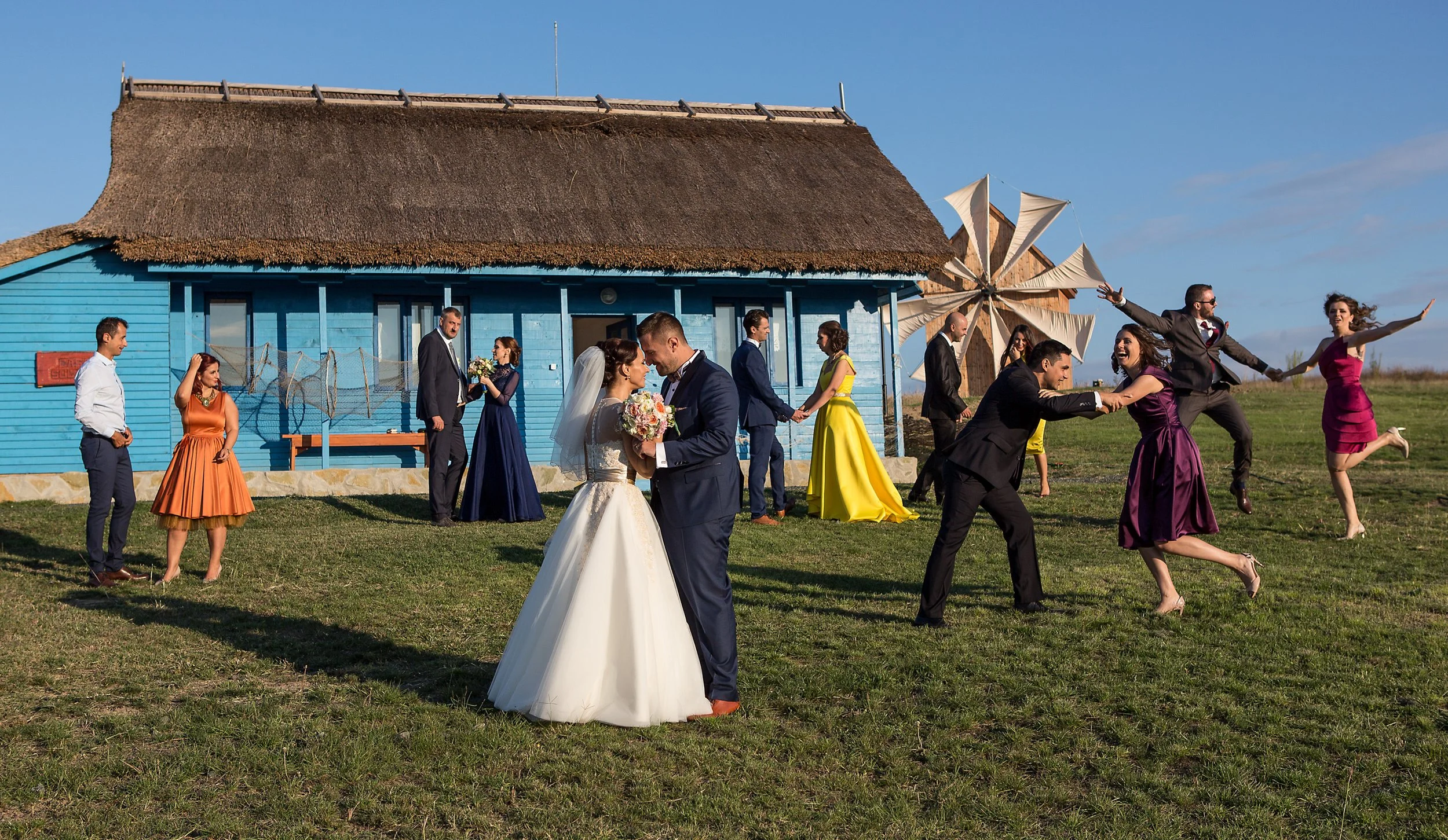 Wedding celebration with bride and groom dancing outdoors near a blue house with a thatched roof, with guests in colorful dresses and suits, and a windmill in the background.