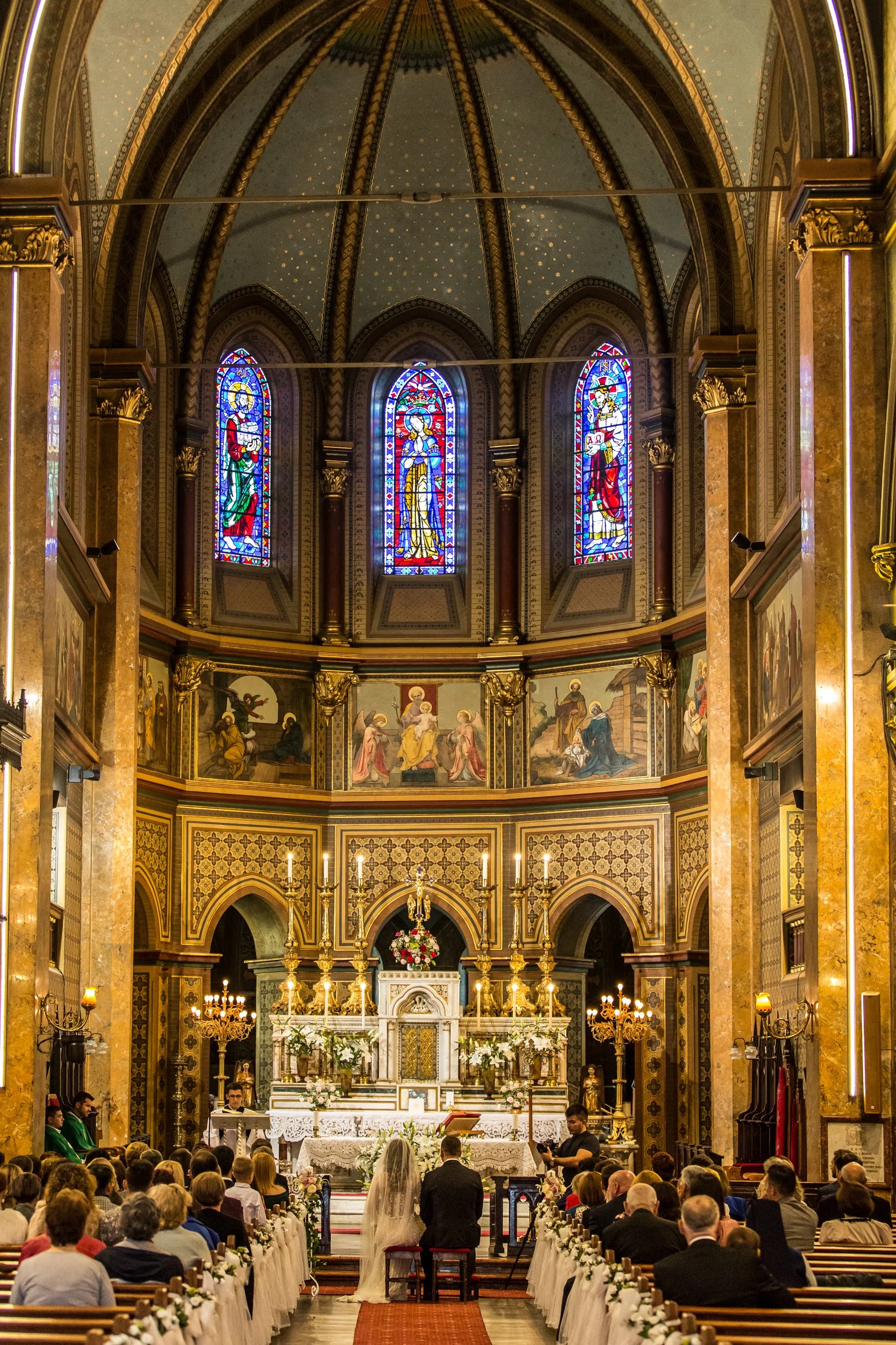 Wedding ceremony inside a church with stained glass windows, an altar decorated with flowers, and many seated guests.