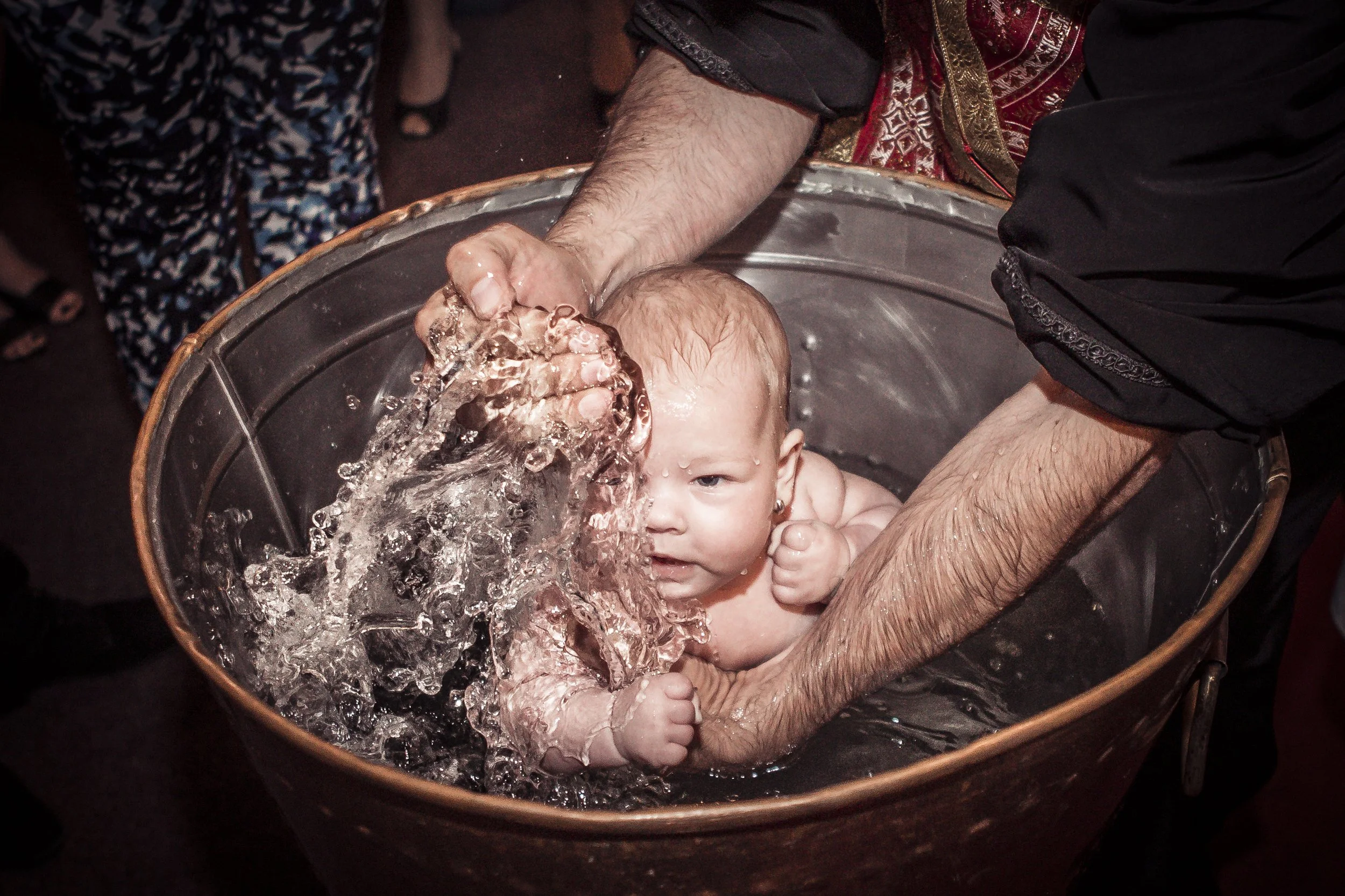 A person is giving a baby a bath in a metal basin, splashing water on the baby's head while holding them securely.