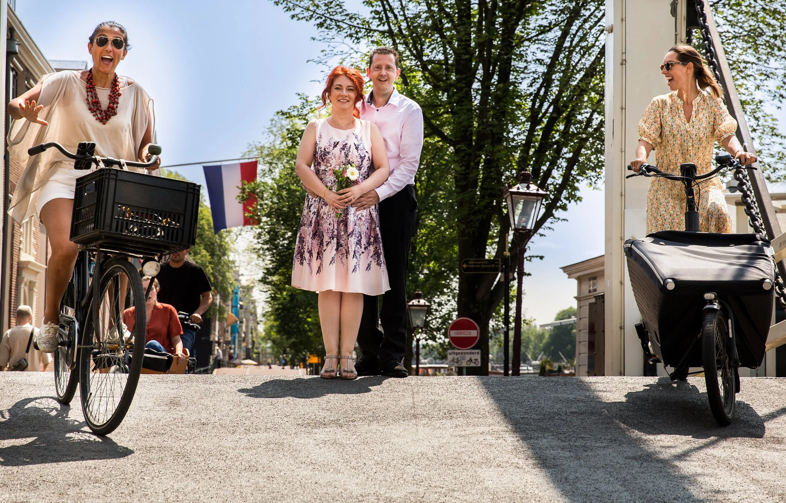 People walking in a city street on a sunny day with trees and historic buildings in the background, two women on bicycles, and a couple walking together.