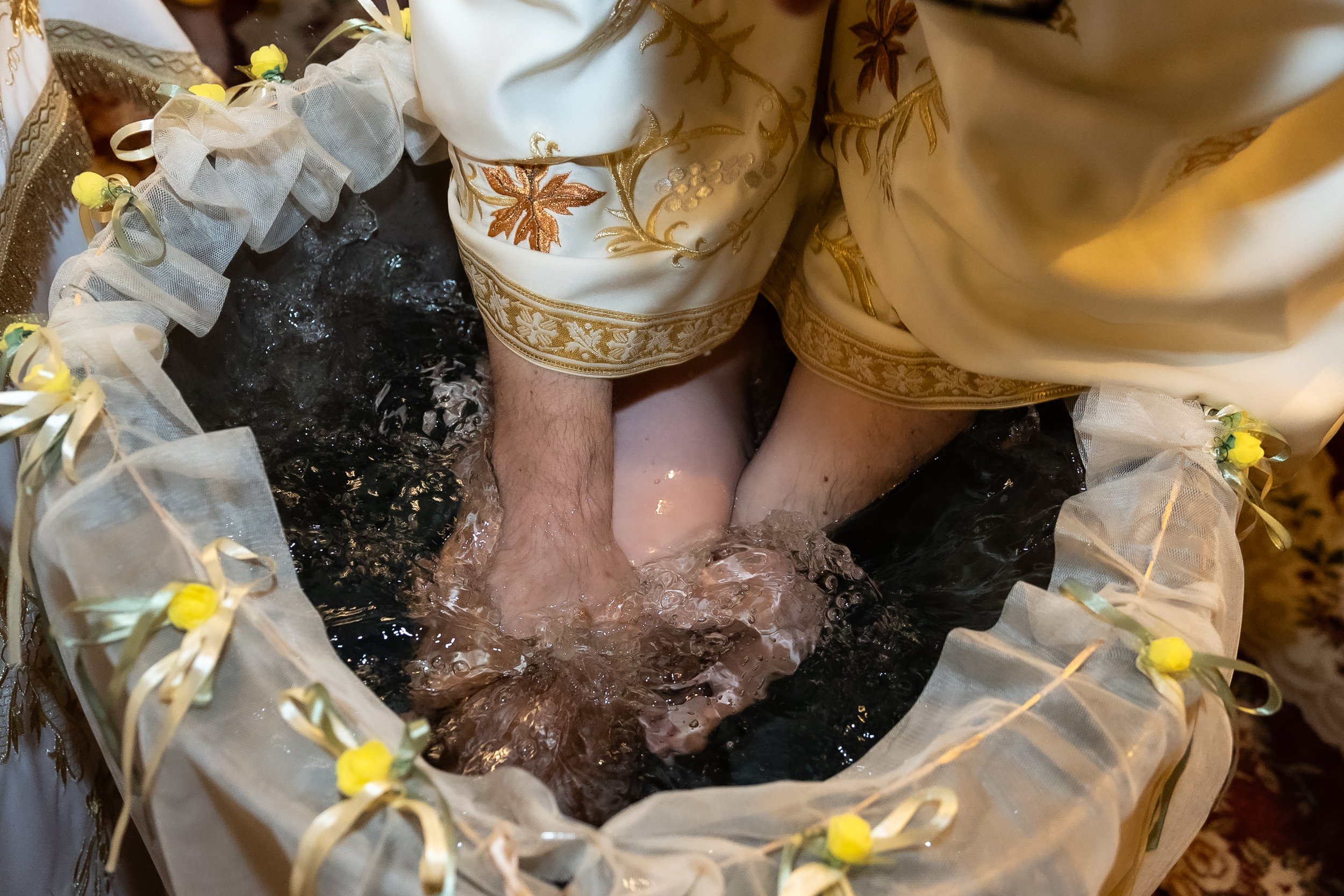 Close-up of a person dressed in ornate, gold-embroidered ceremonial clothing, sitting in a black basin filled with water, surrounded by decorative items and soft ribbons with small yellow flowers.