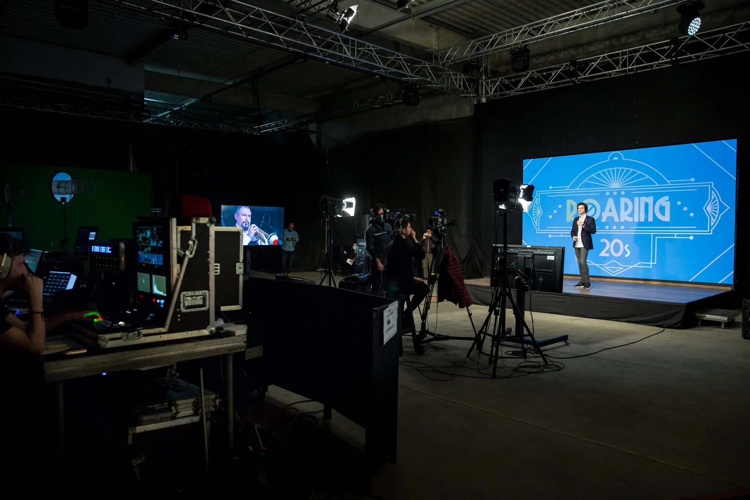 A person is standing on a stage delivering a presentation at an event called 'ROARING 20s'. There are cameras and crew members capturing the event in a dark indoor setting. A large blue screen with event branding is visible behind the speaker.