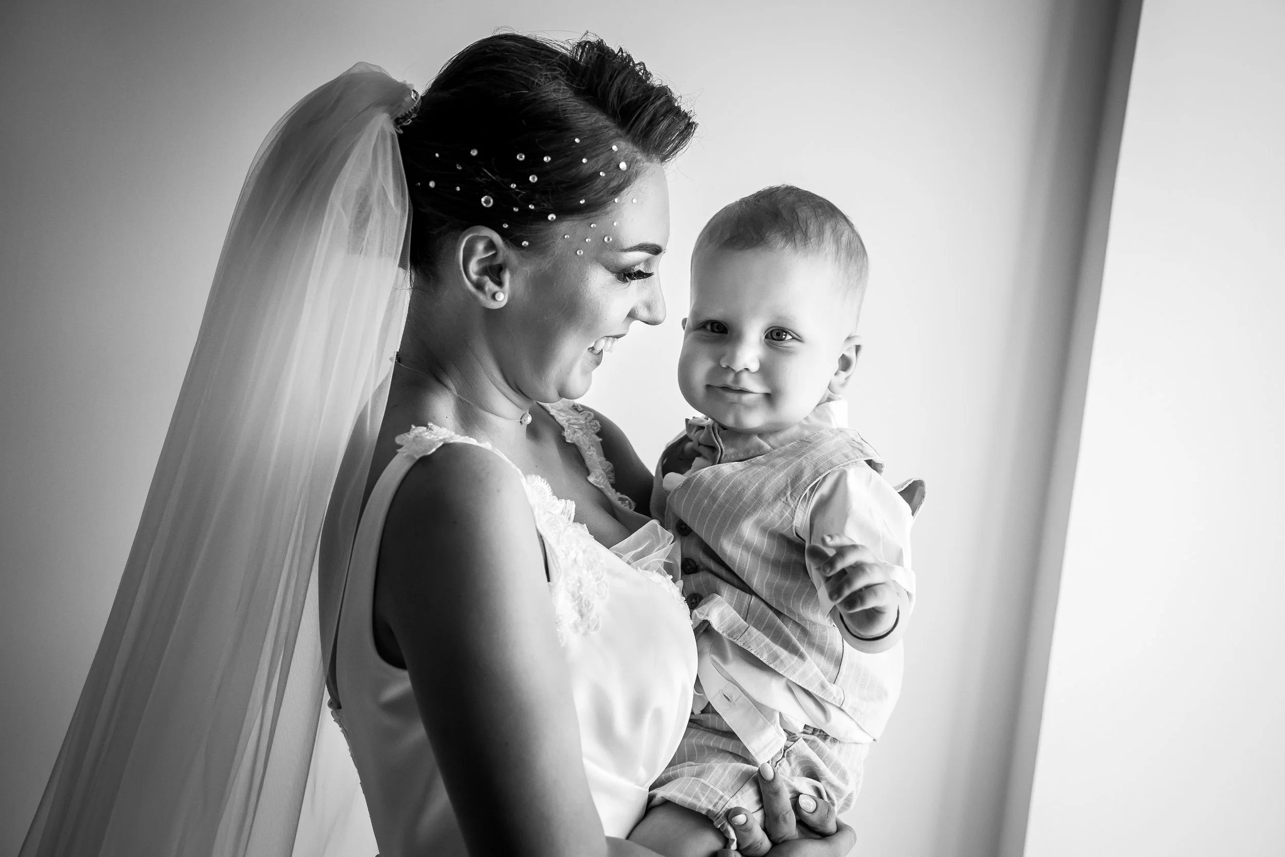 Black and white photo of a woman in a wedding dress holding a young boy, both smiling and looking at each other.