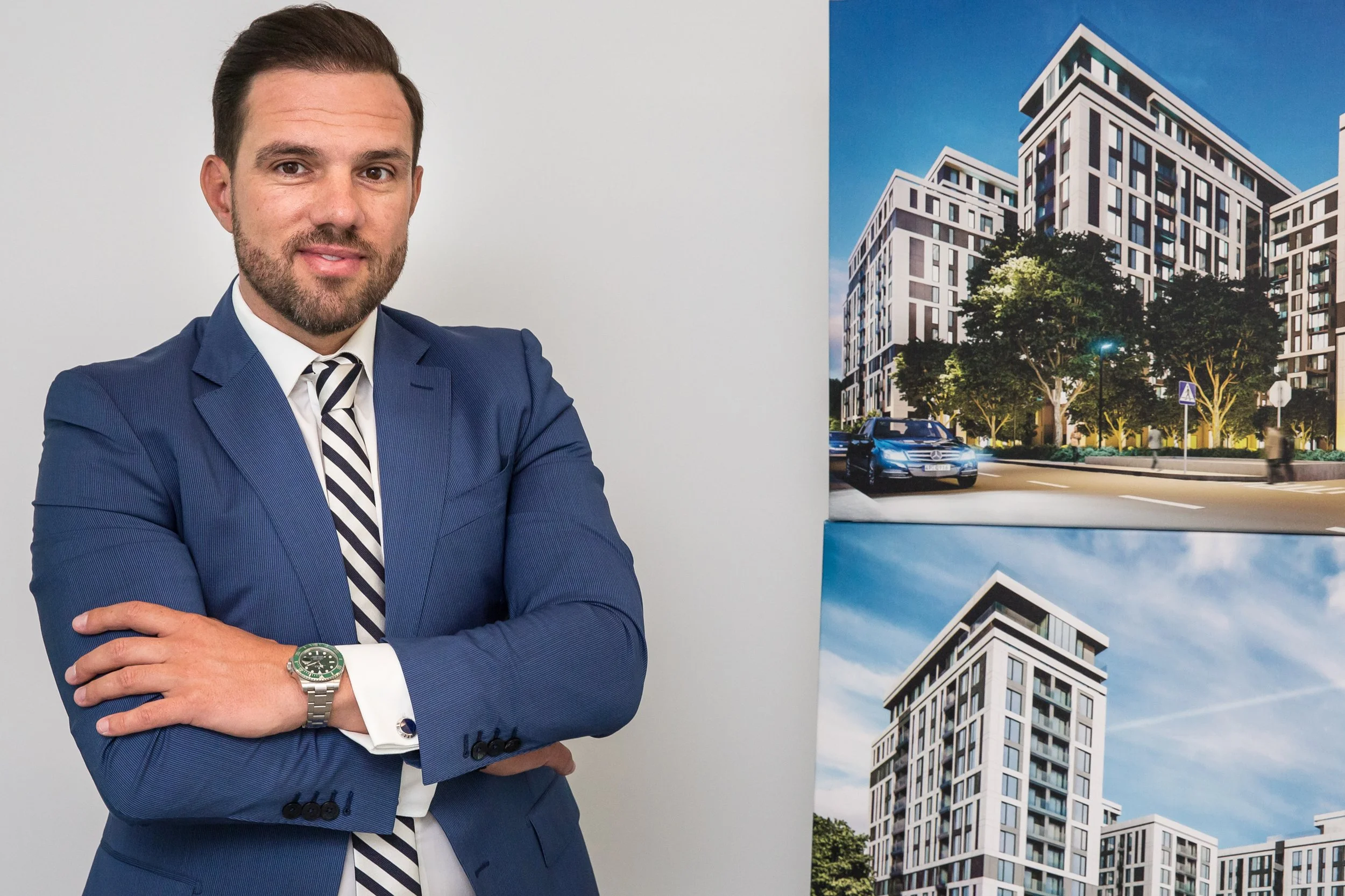 A man dressed in a blue suit with a white shirt and striped tie standing with his arms crossed, wearing a silver and green watch, standing in front of architectural displays of modern multi-story buildings and cityscapes.