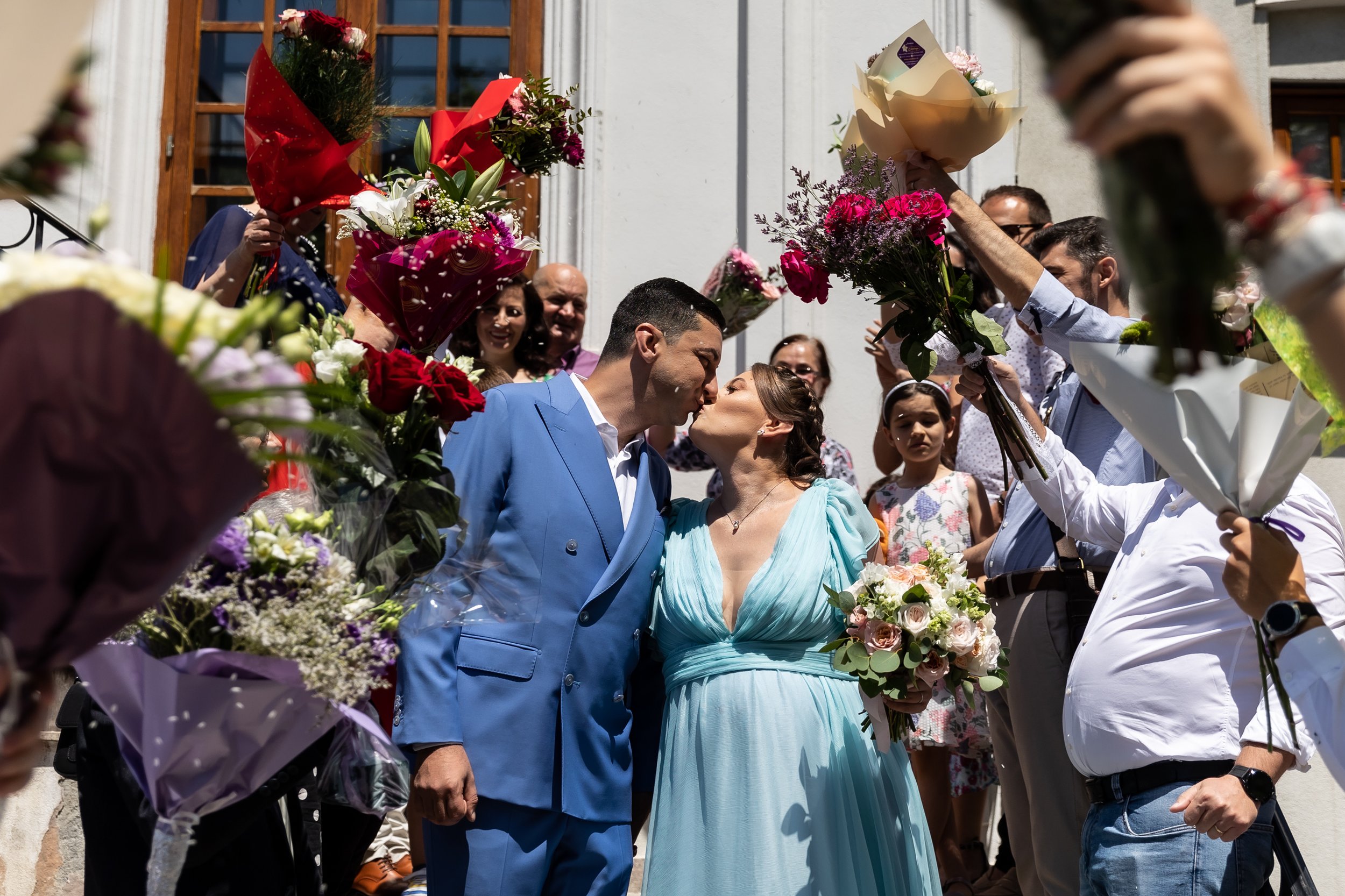 A wedding scene with a couple in the center sharing a kiss, surrounded by people holding bouquets of flowers and reaching out to celebrate them.