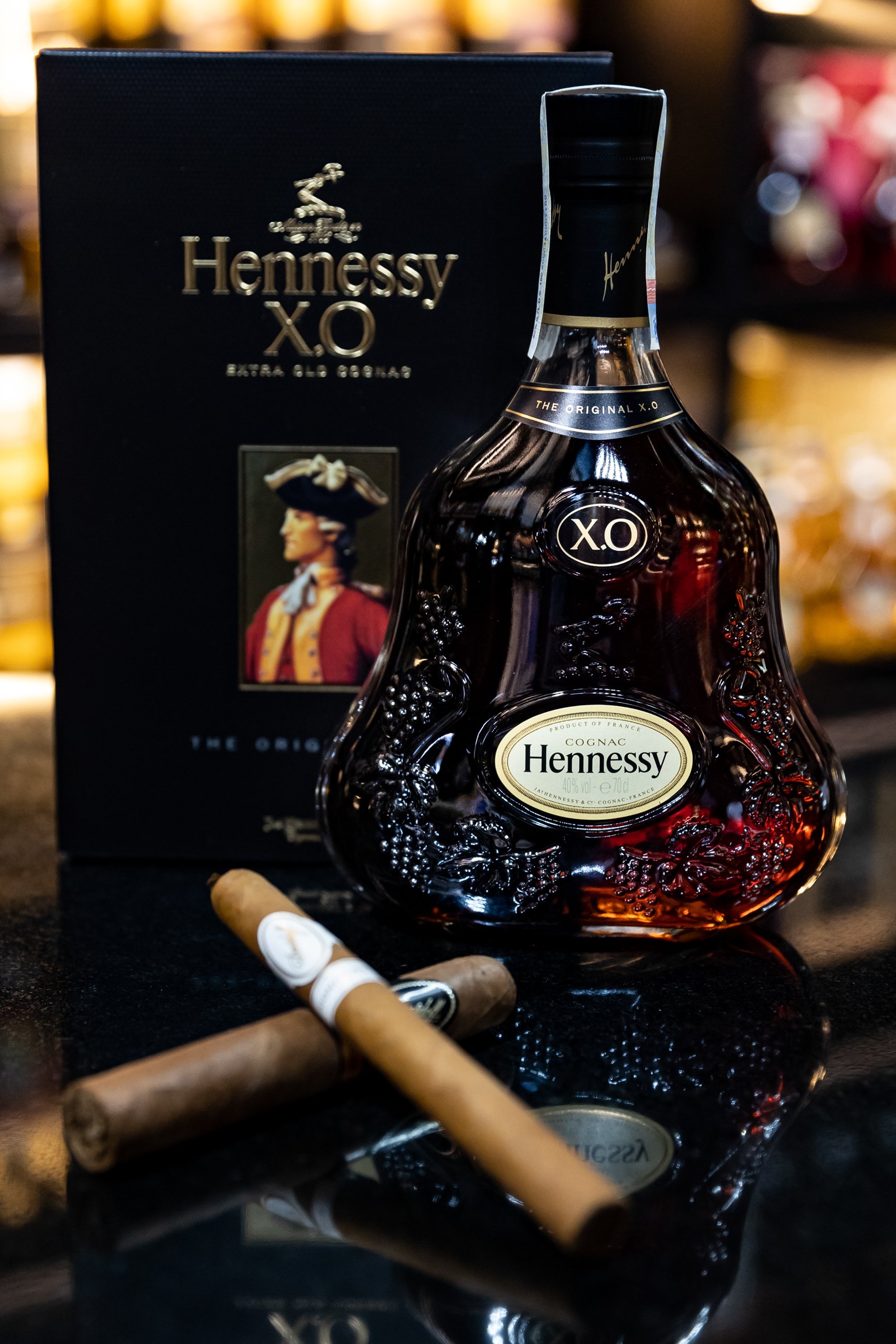 A bottle of Hennessy X.O cognac with its box behind it, placed on a black surface. Two cigars are in the foreground, and the background is blurred with warm lighting.