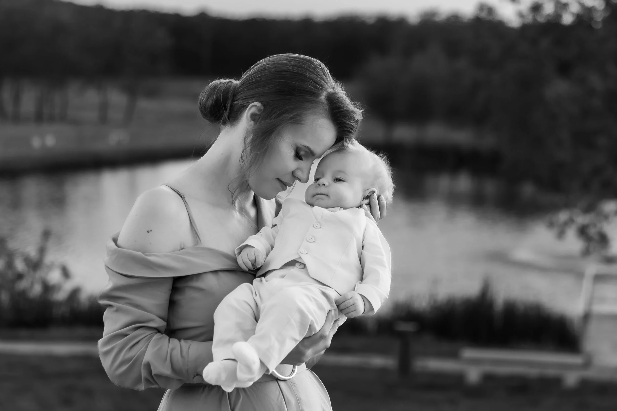 A woman holding a baby outdoors near a body of water, with a blurred background of trees and landscape, black and white photograph.