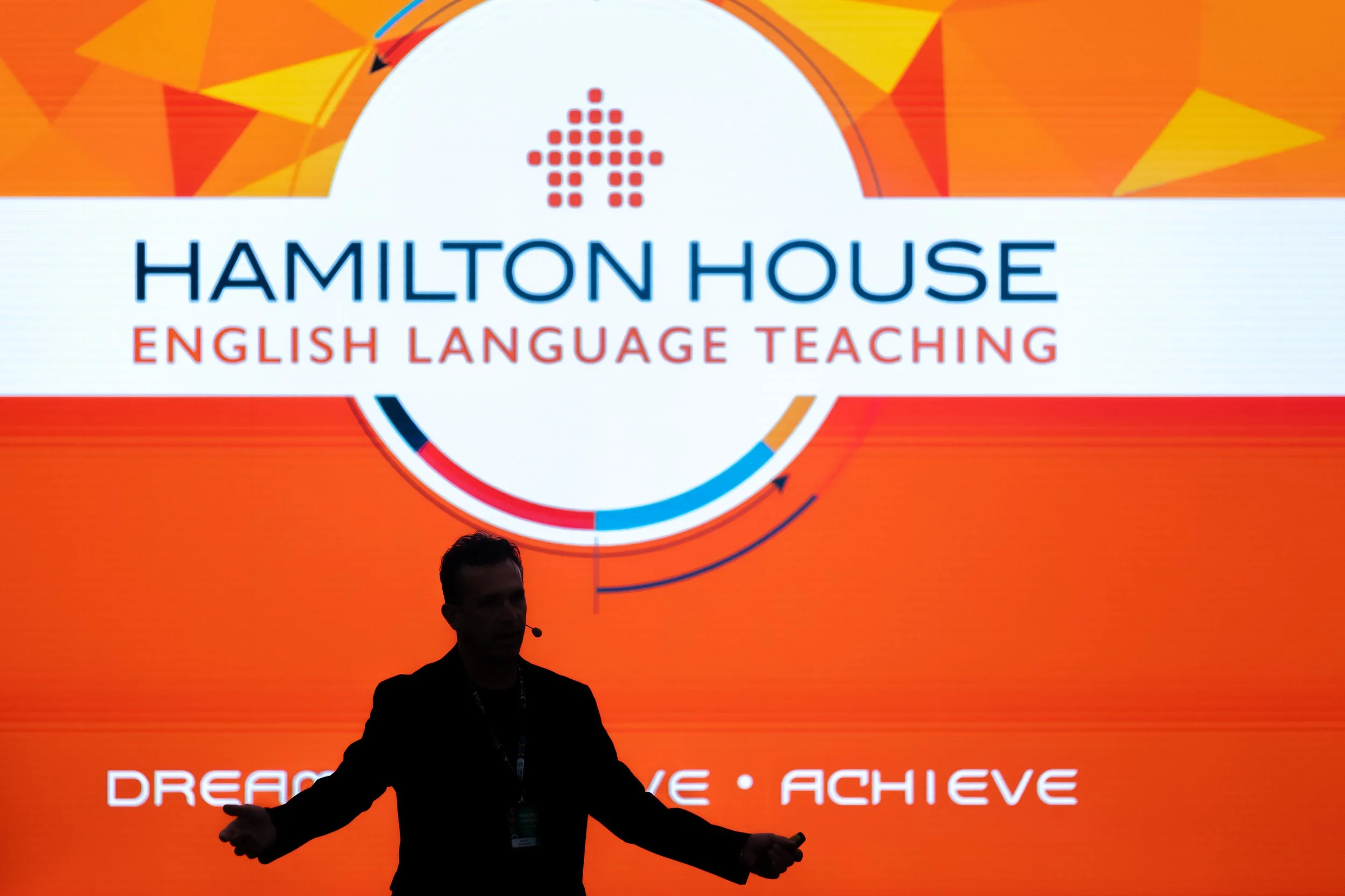 A person is speaking at a presentation with a large orange and red geometric background. The slide behind features a logo with a dotted upward arrow and the words 'HAMILTON HOUSE' and 'ENGLISH LANGUAGE TEACHING' in blue and red text, respectively. Th
