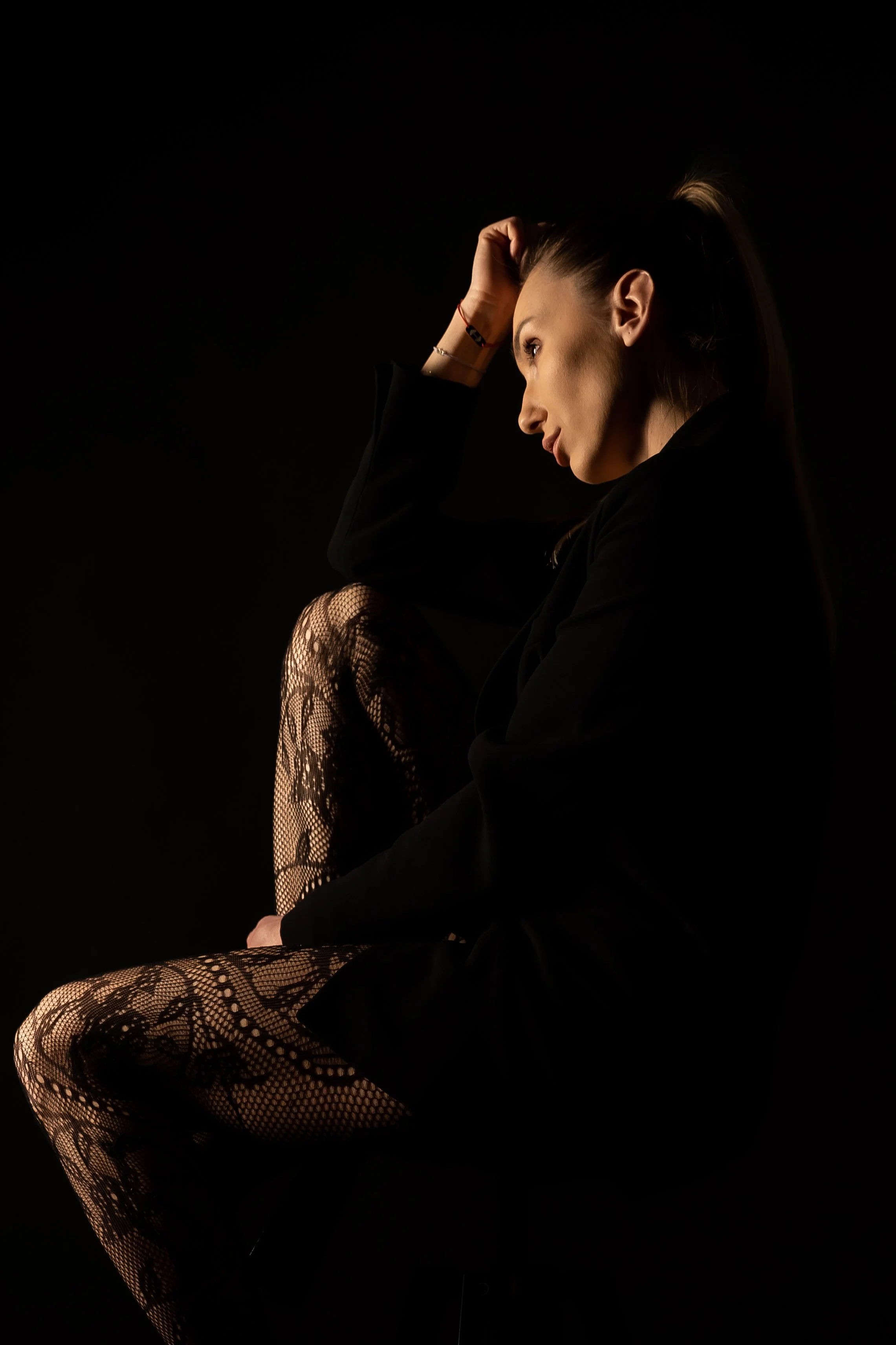 A woman with a ponytail, wearing a black blazer, sitting with one knee raised and her head resting on her hand, on a black background. She is wearing lace tights.